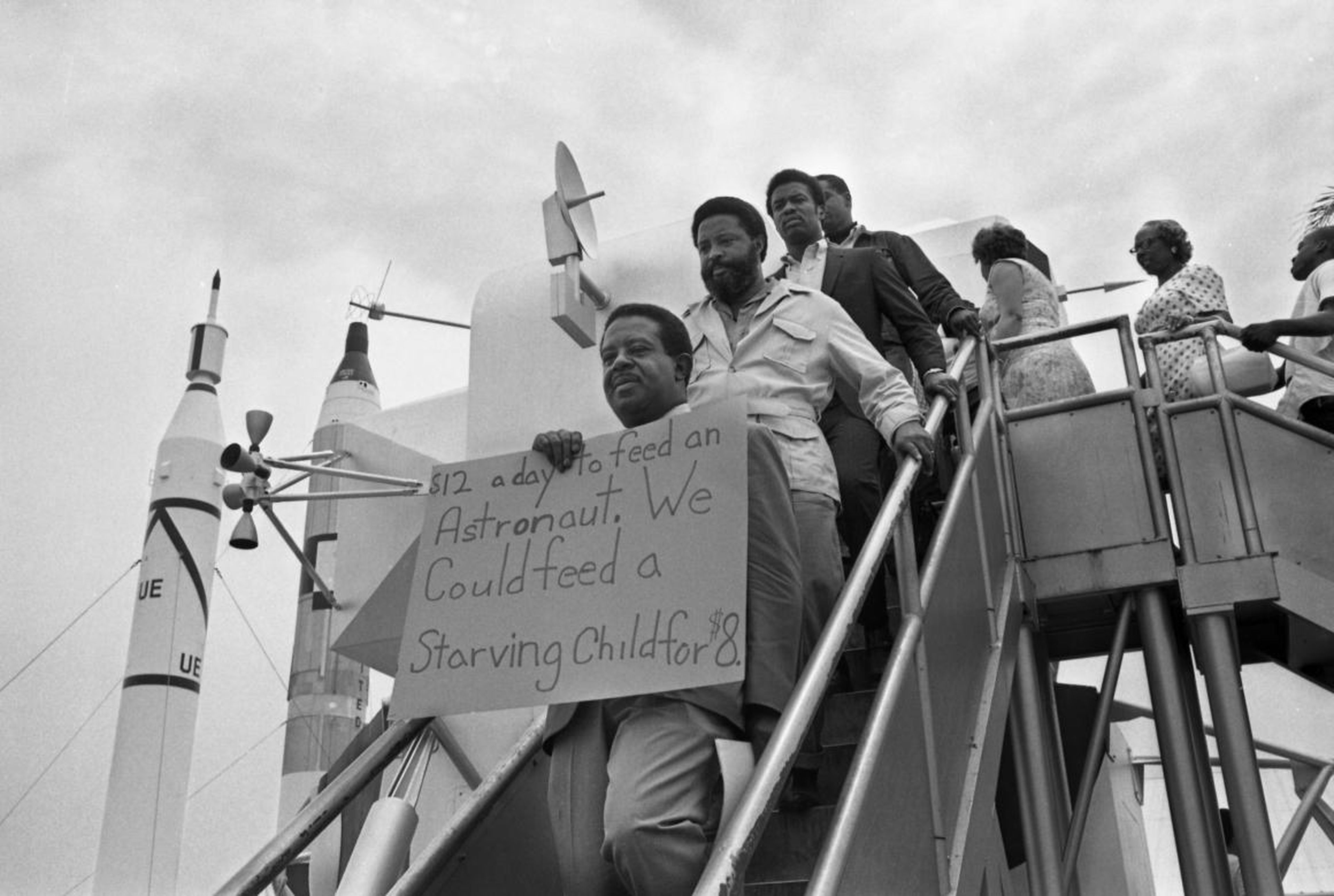 El reverendo Ralph Abernathy se para en los escalones de una maqueta del módulo lunar que muestra un cartel de protesta mientras se manifiesta en el lanzamiento de la luna del Apolo 11.