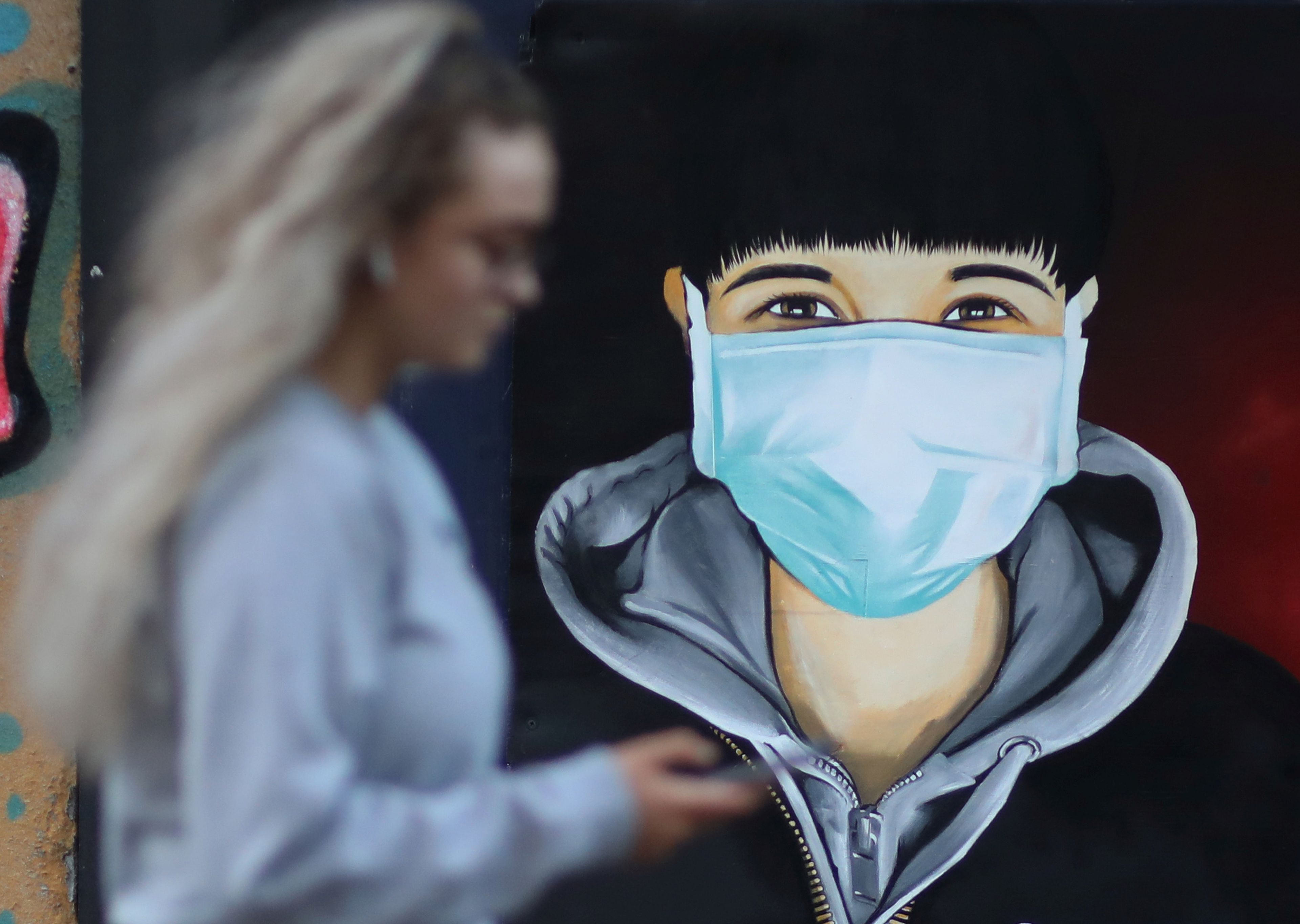 Una mujer mira su móvil mientras camina junto a un mural con un hombre llevando mascarilla.