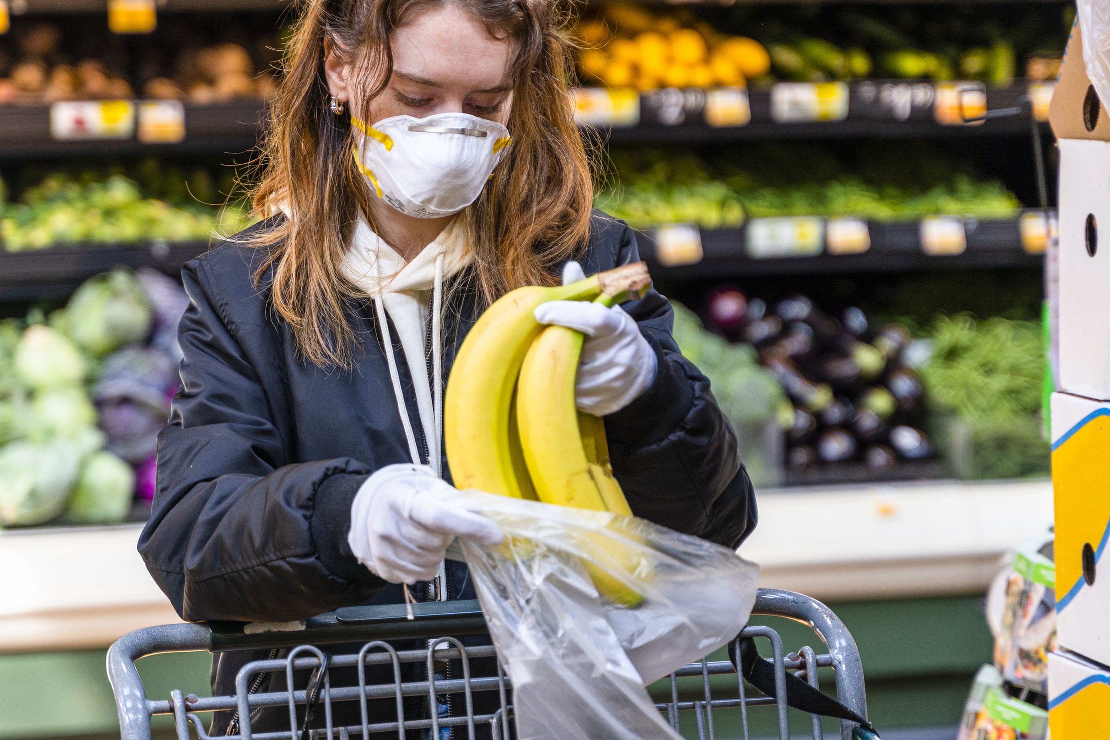 Chica comprando plátanos en un supermercado con mascarilla y guantes.
