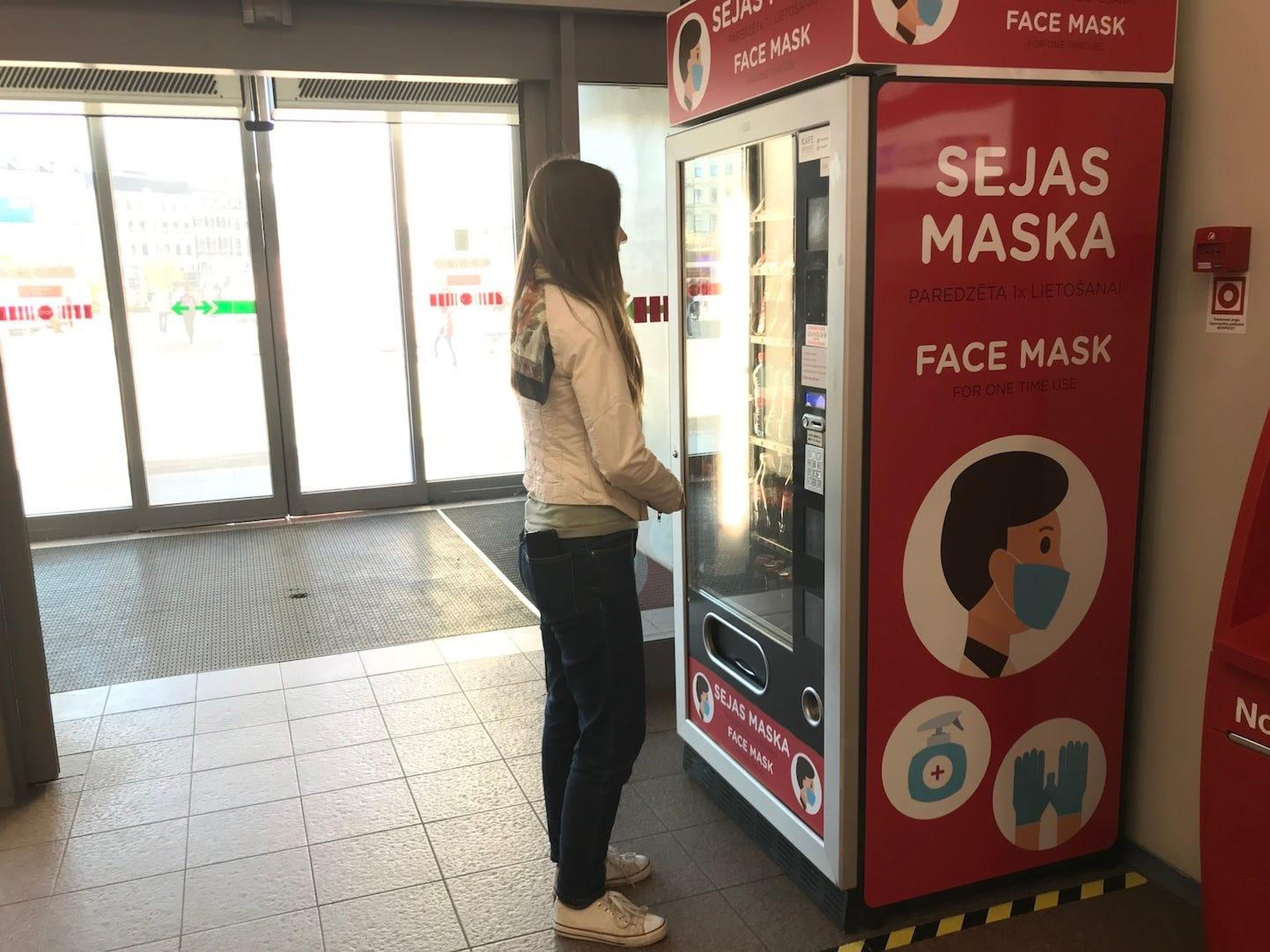 Una mujer en frente de una máquina expendedora de mascarillas desechables en una estación de tren de Riga.