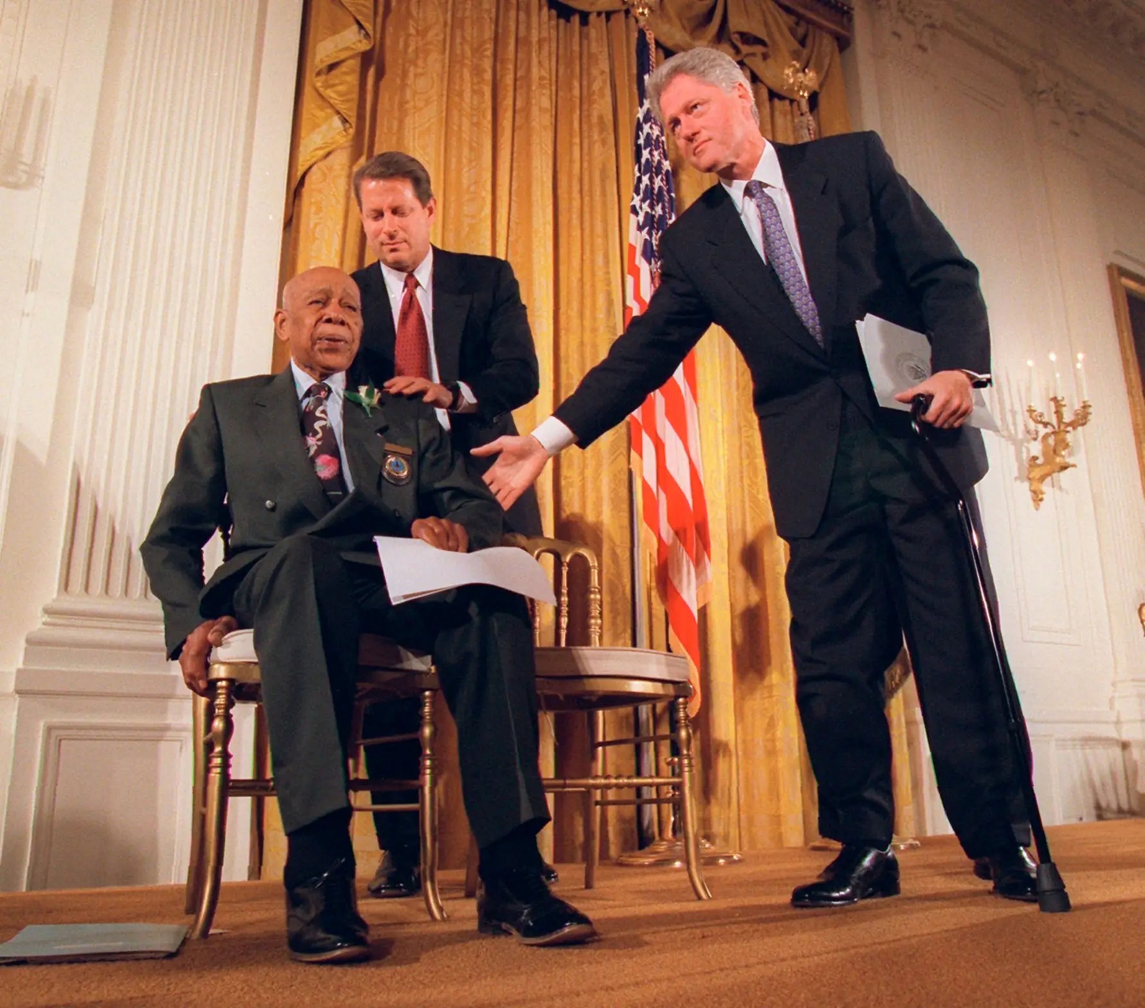 El presidente Bill Clinton y el vicepresidente Al Gore ayudan a Herman Shaw, de 94 años, sobreviviente del experimento de Tuskegee, durante una conferencia de prensa el 16 de mayo de 1997.