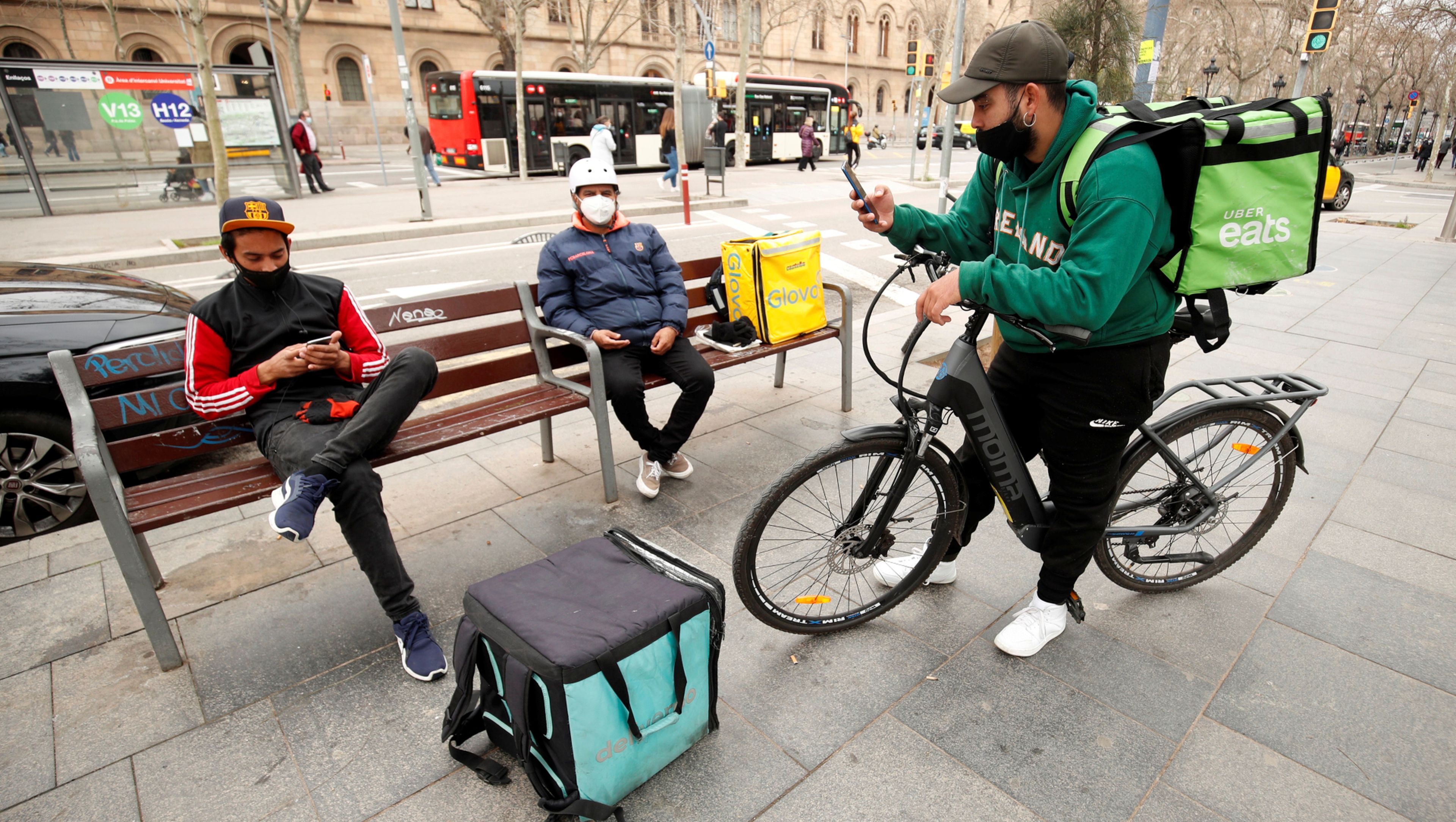 Riders de Glovo, Deliveroo y Uber Eats esperan pedidos en un banco en Barcelona
