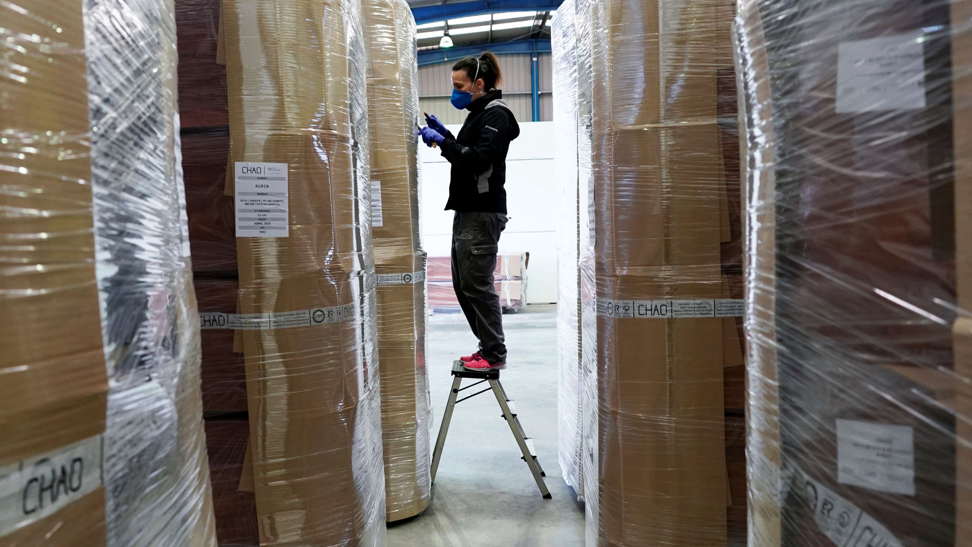 Una trabajadora con mascarilla revisa cajas de ataúdes en un almacén logístico
