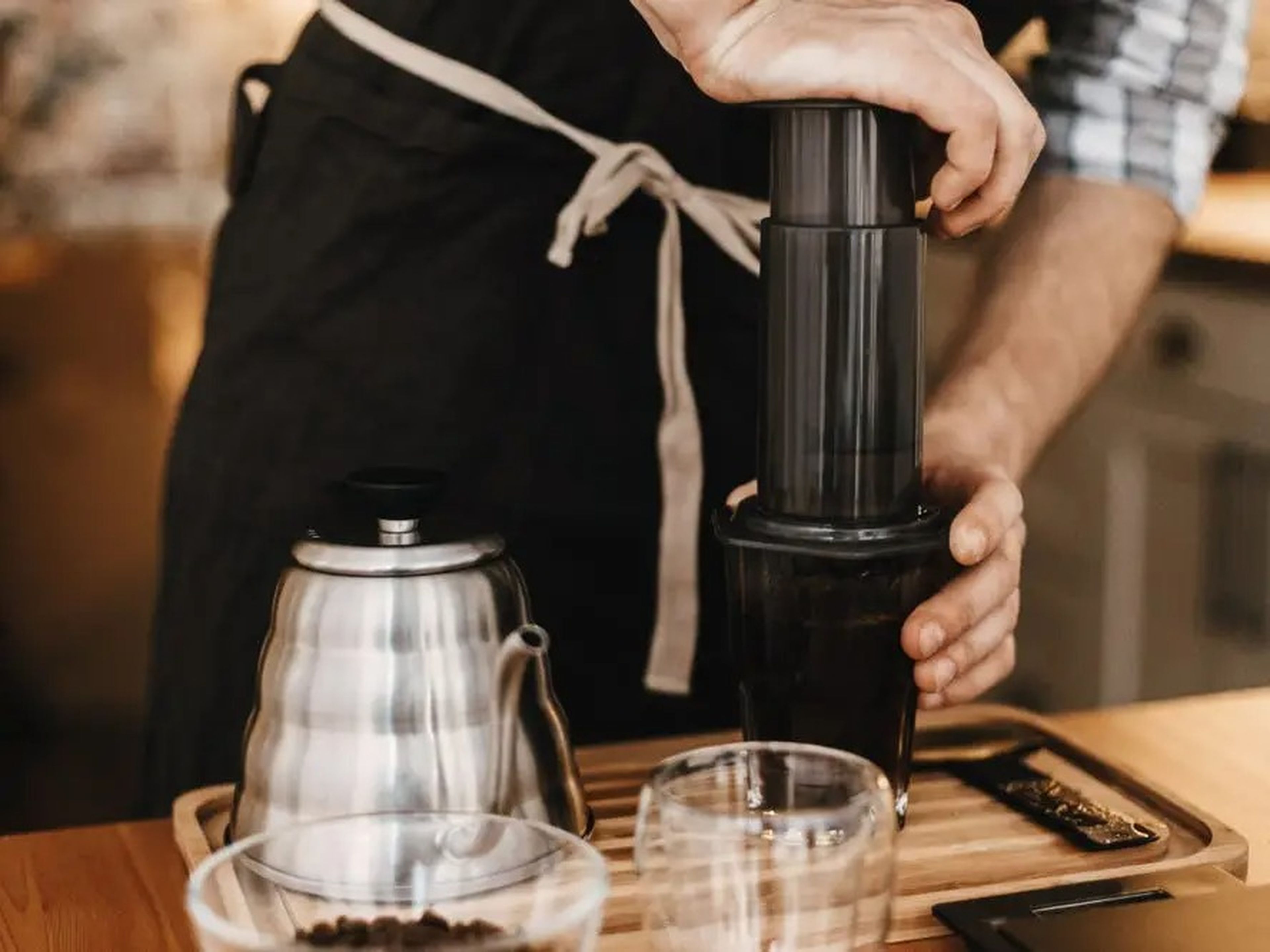 Professional barista preparing coffee by AeroPress and glass cup, scales, manual grinder, coffee beans, kettle on wooden table