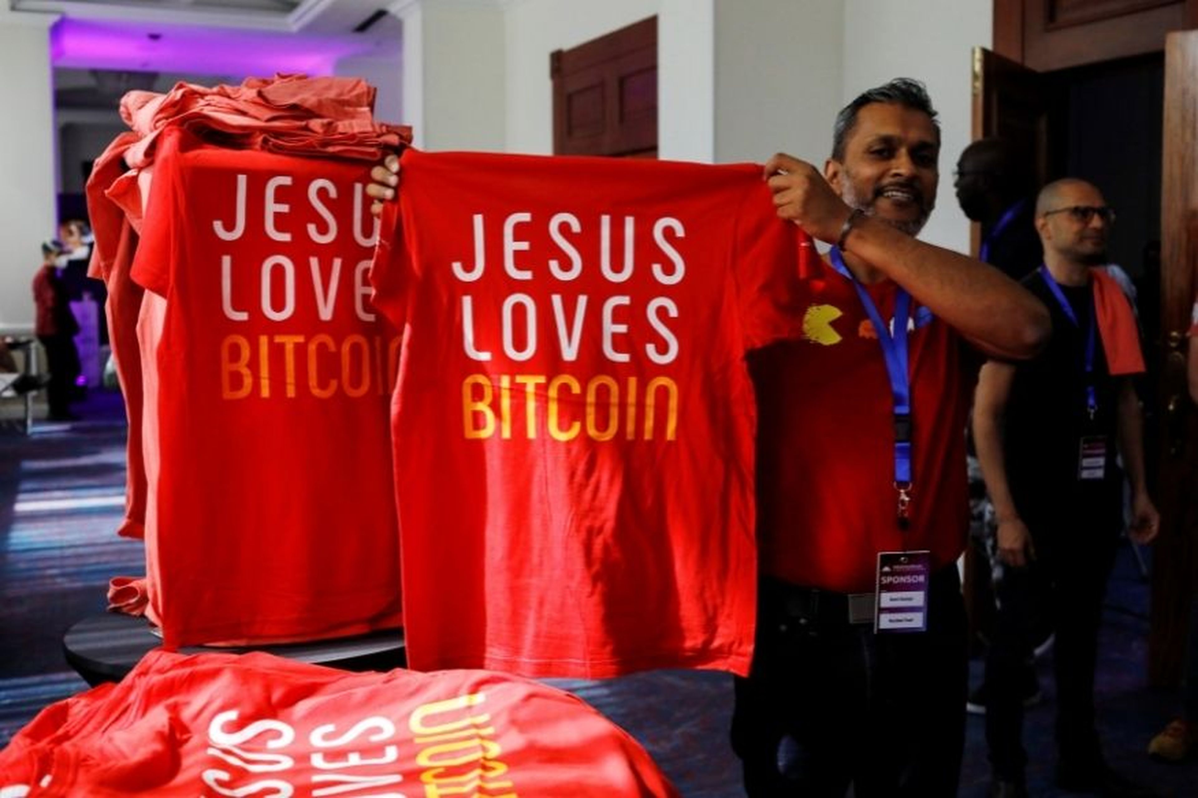 Un hombre muestra unas camisetas en las que se lee: “Jesús ama bitcoin”.