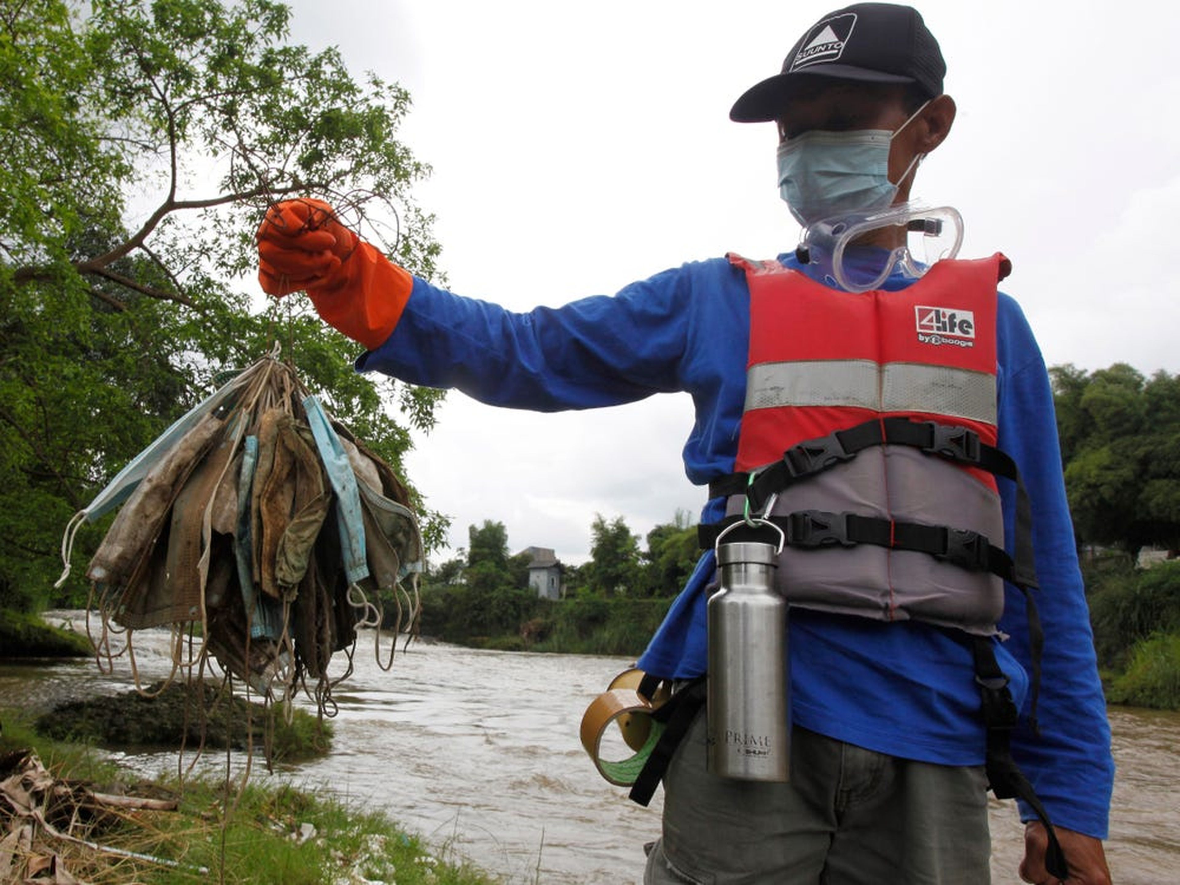 Un hombre con chaleco salvavidas y guantes sostiene una colección de máscaras sucias frente a un río.
