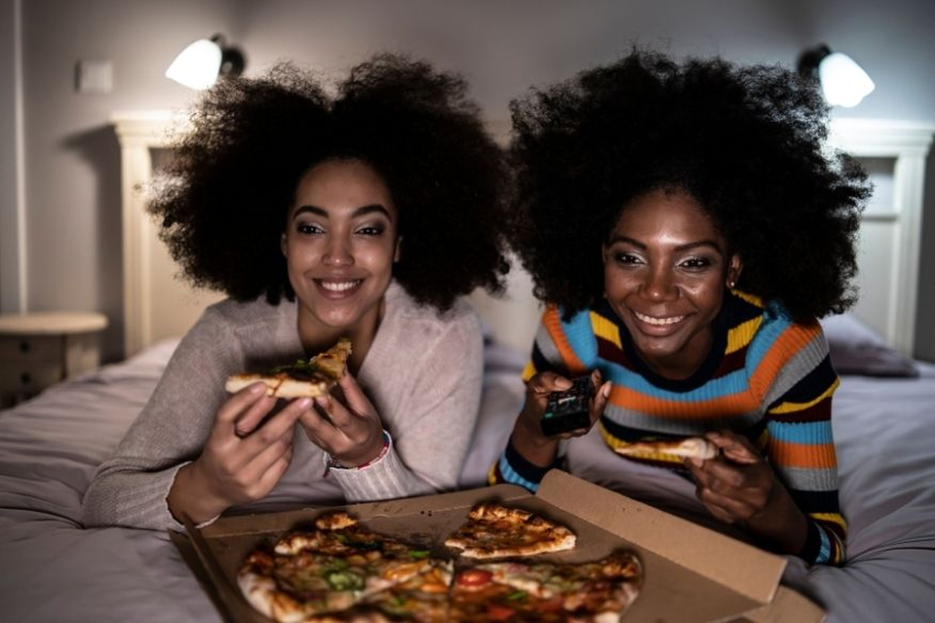 2 mujeres comen pizza encima de la cama.