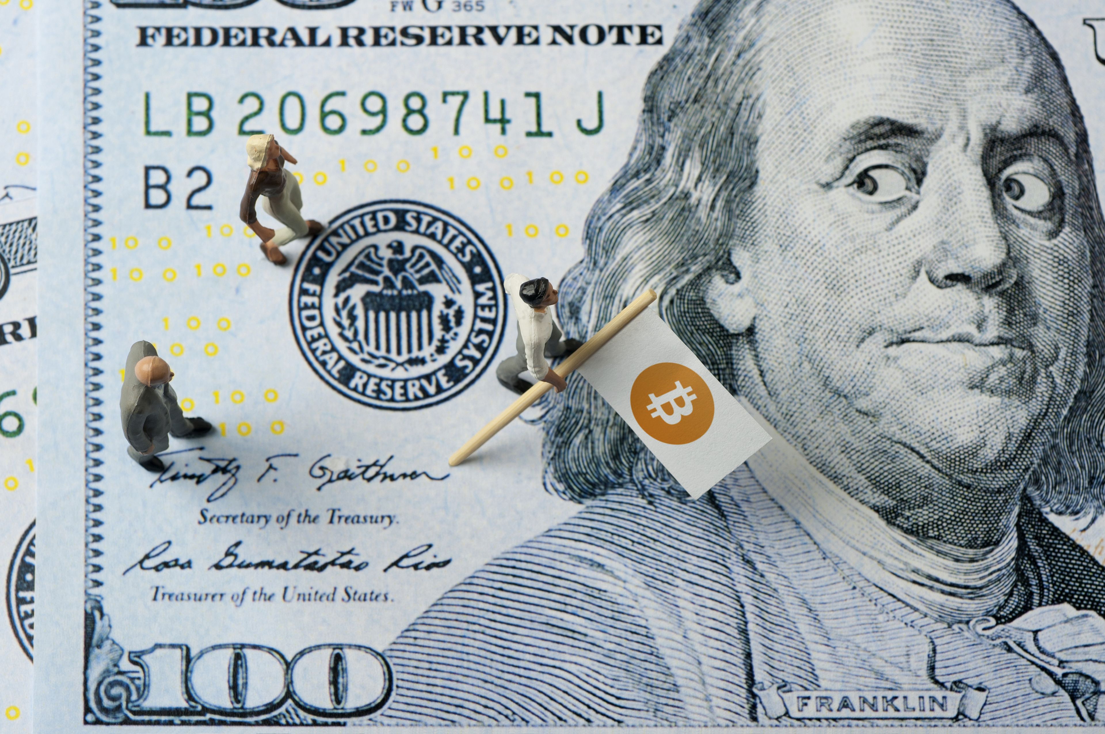Un muñeco coloca una bandera de Bitcoin en un billete de 100 dólares.