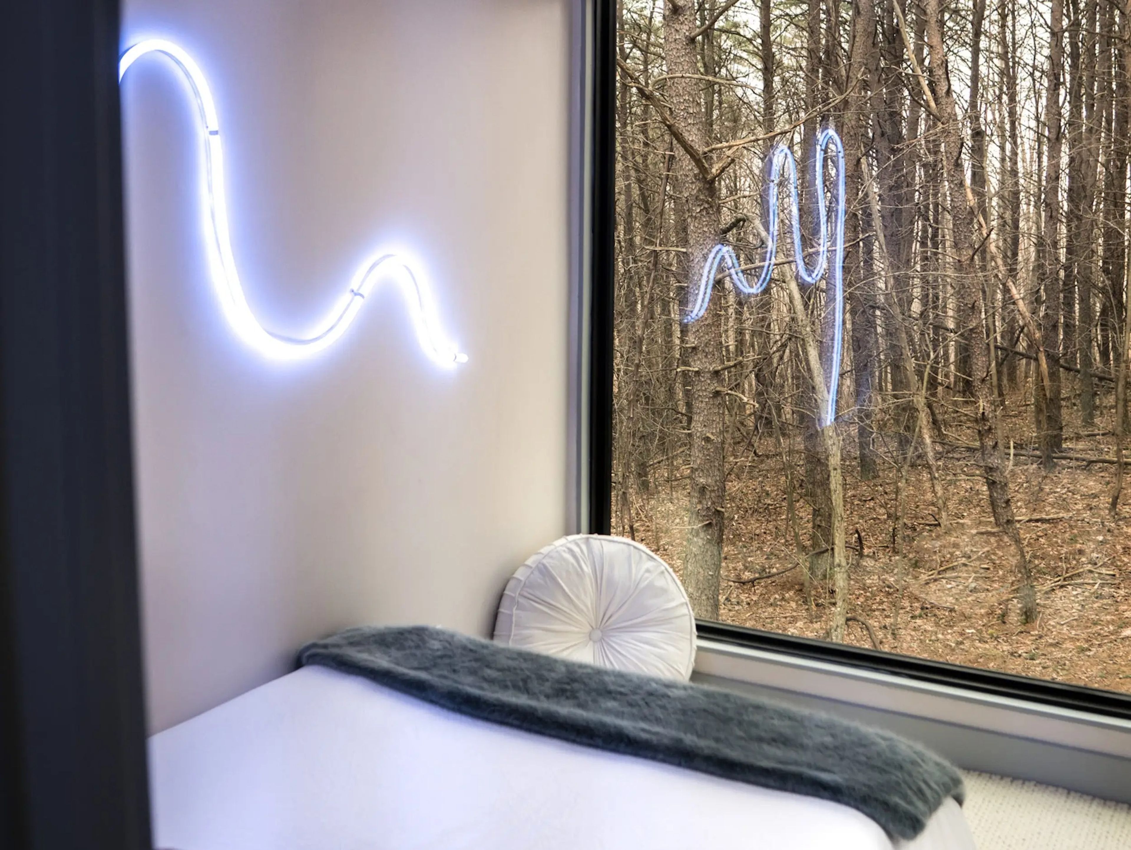 Wavy neon lights next to a bed overlooking a window of walls with views of the tree inside the OG Box, a shipping container home.
