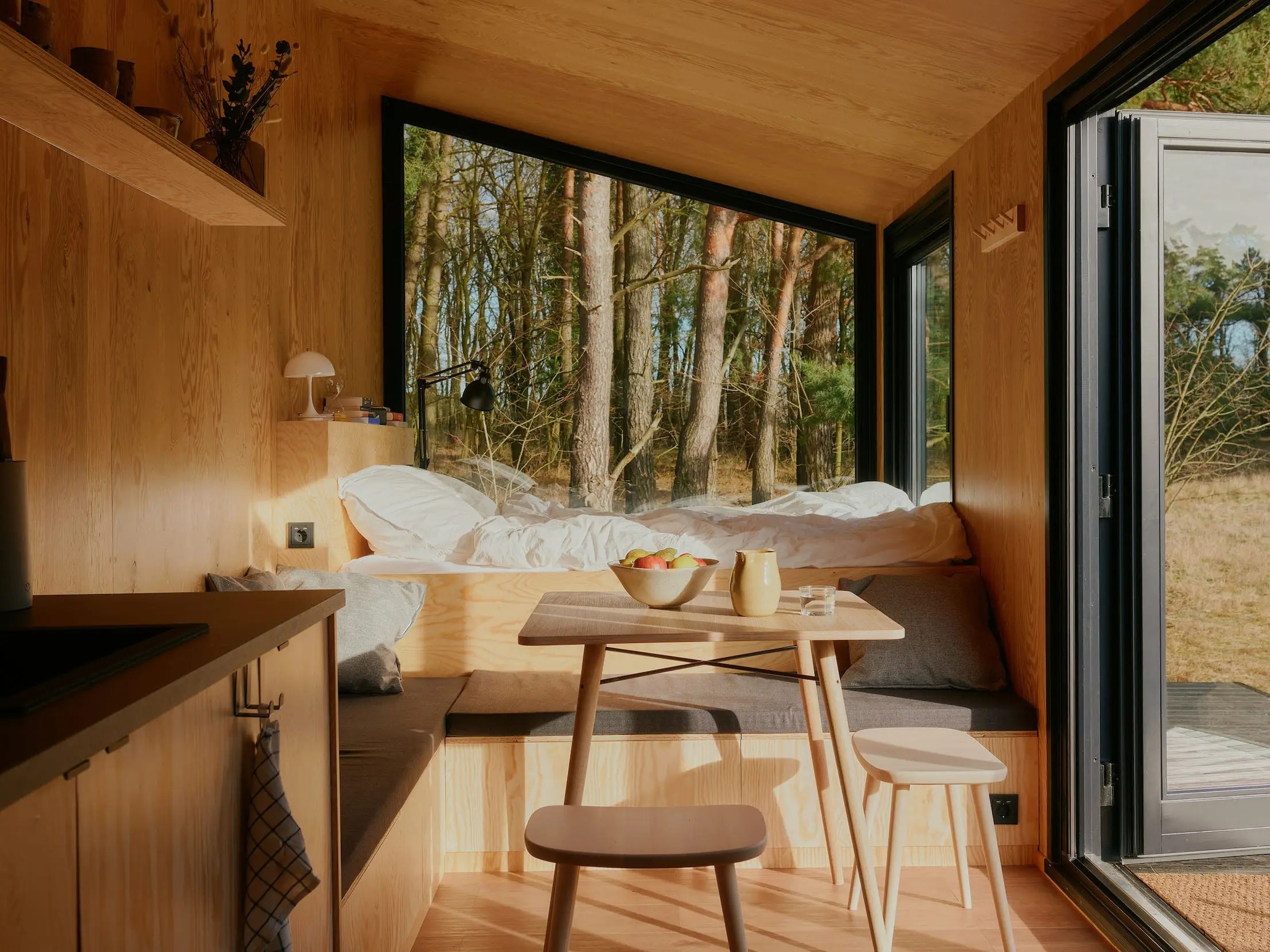 Una cama, una cocina, una mesa con sillas y ventanas que muestran la naturaleza en el exterior.
