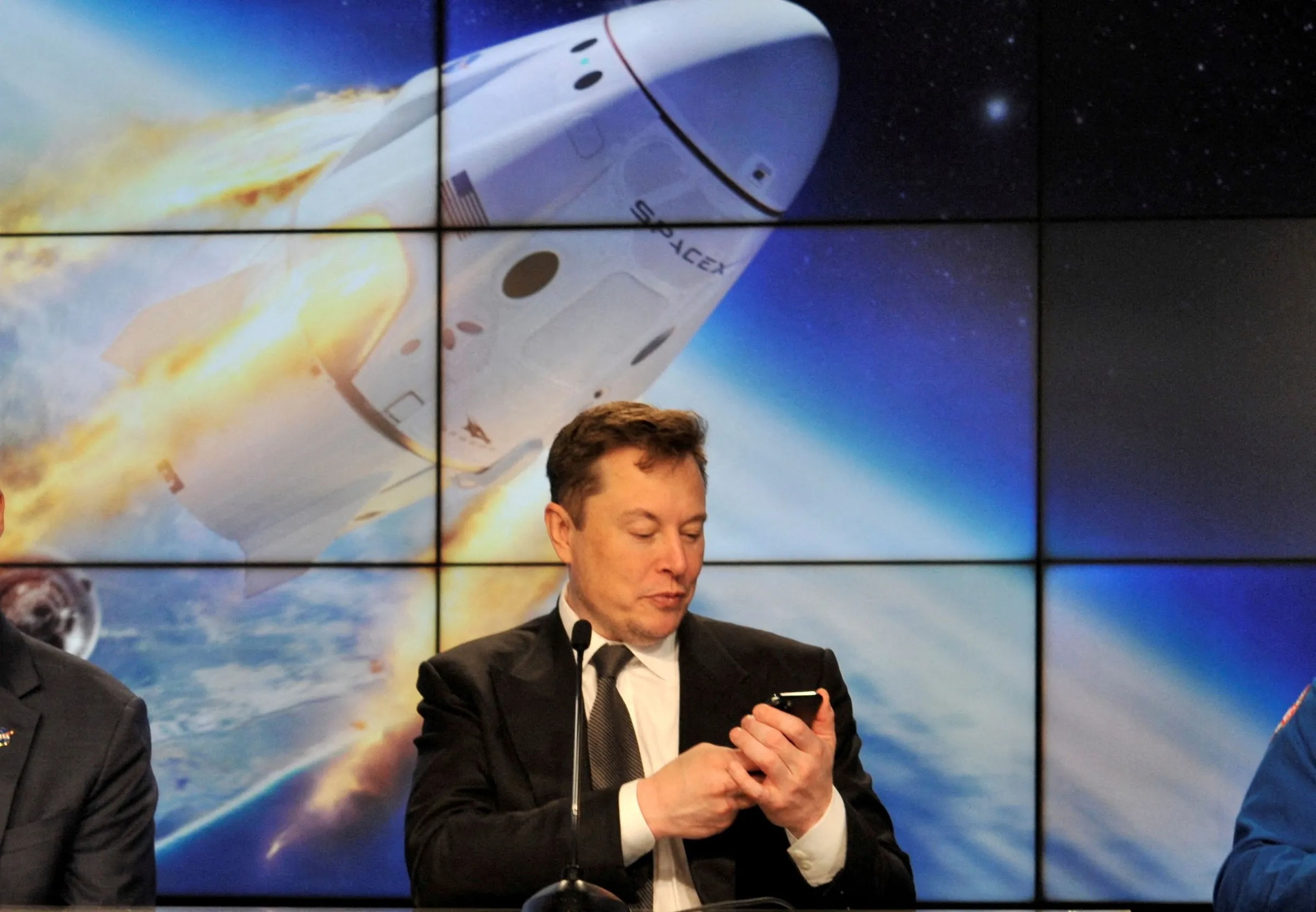 Elon Musk mira su teléfono con una foto de una cápsula espacial entrando en órbita detrás de él.