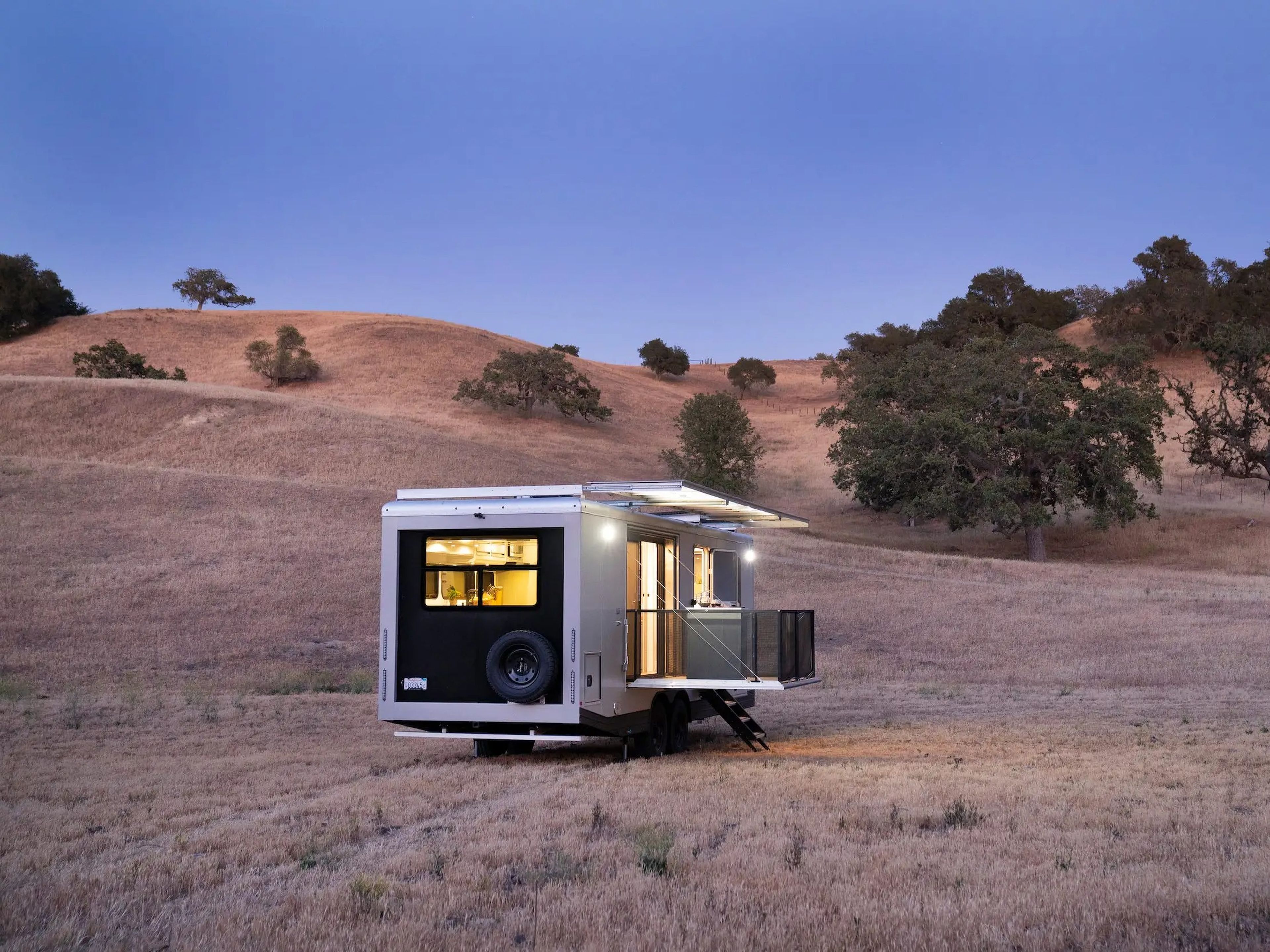 The exterior of the travel trailer as it sits on a brown field at dusk. The lights are on inside.