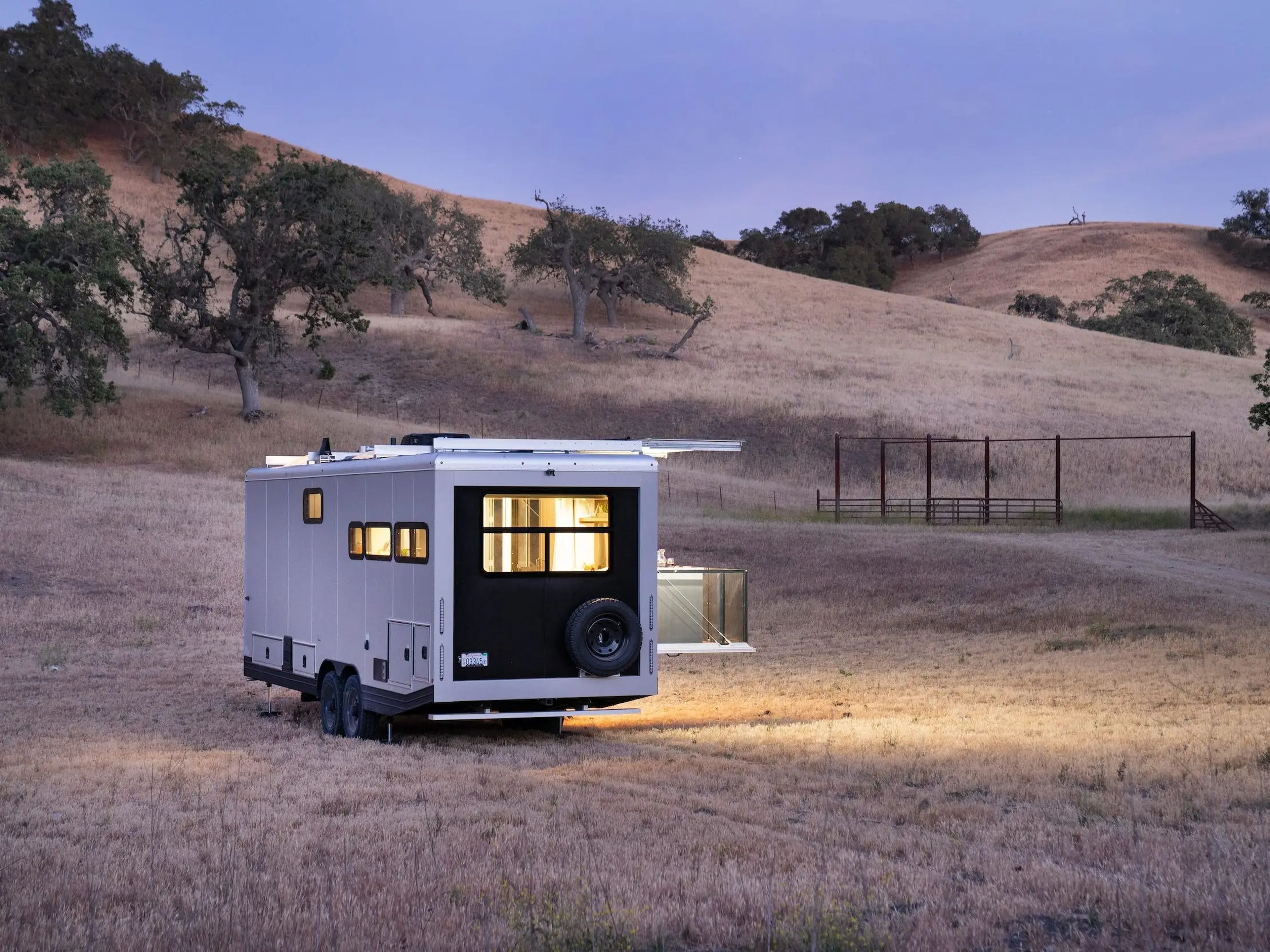 The exterior of the travel trailer as it sits on a brown field at dusk. The lights are on inside.