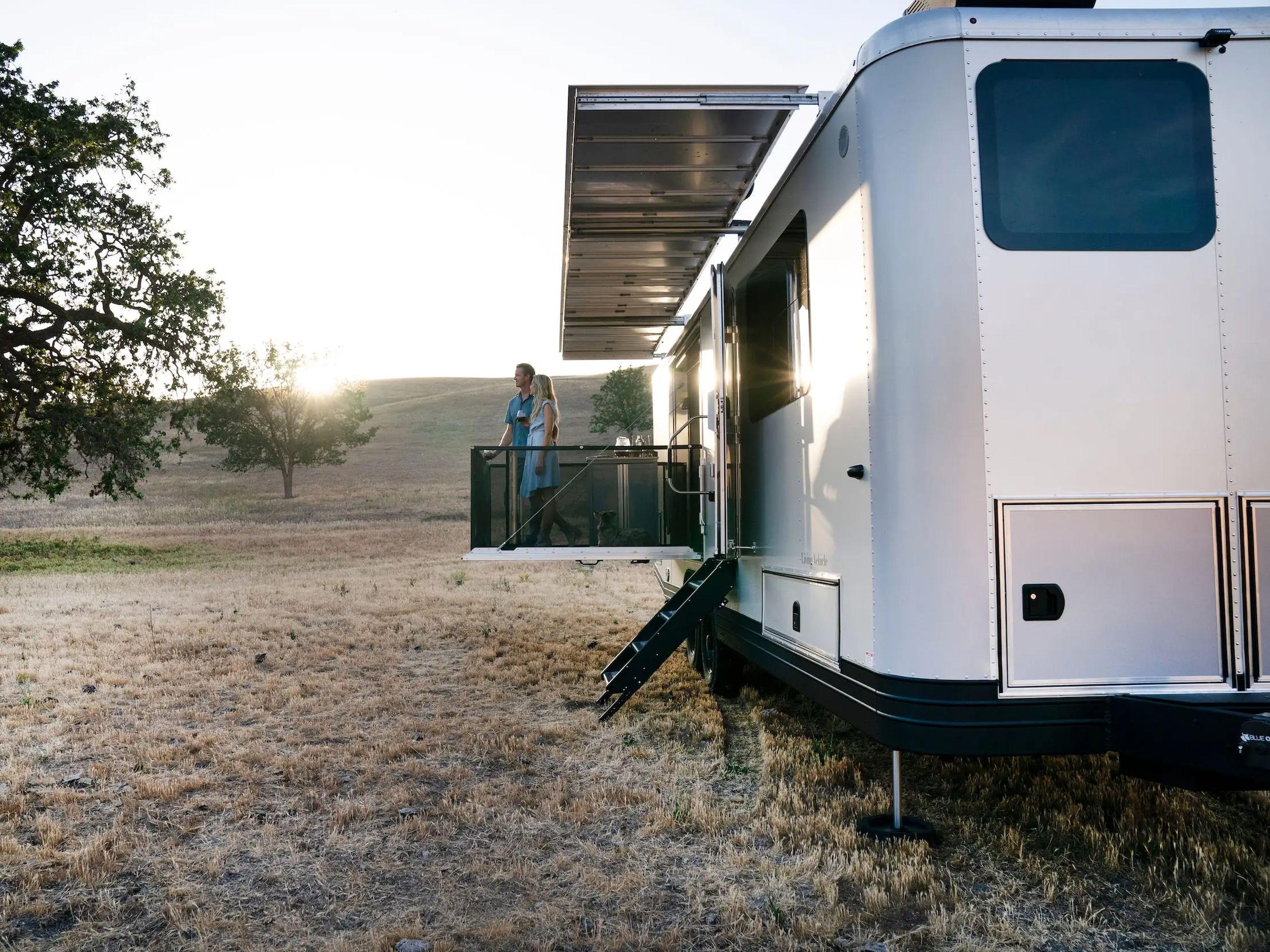 The exterior of the travel trailer as it sits on a brown field. The patio is extended and people are standing on it.