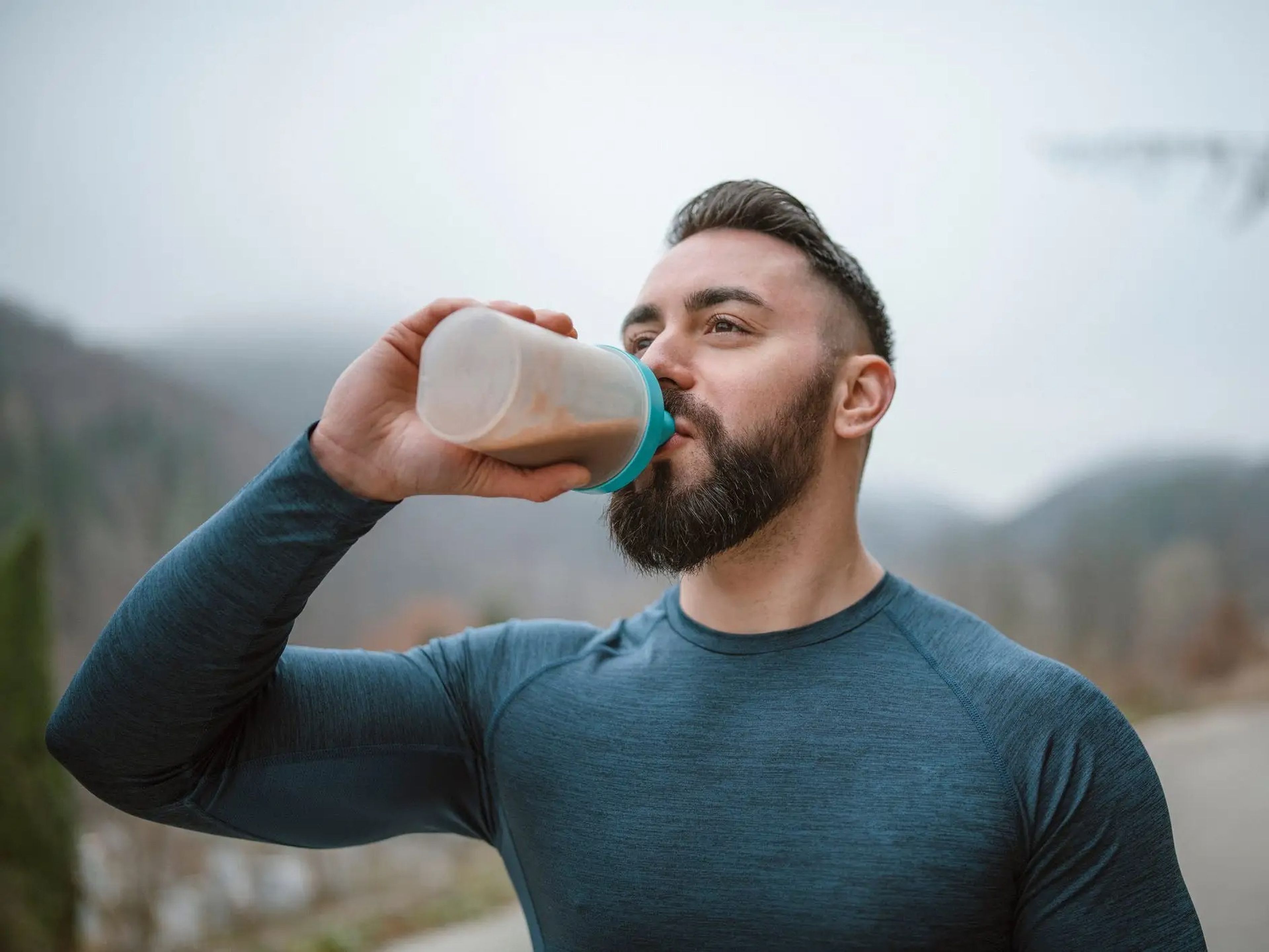 an athletic man in a long sleeved shirt drinking a protein shake outdoors
