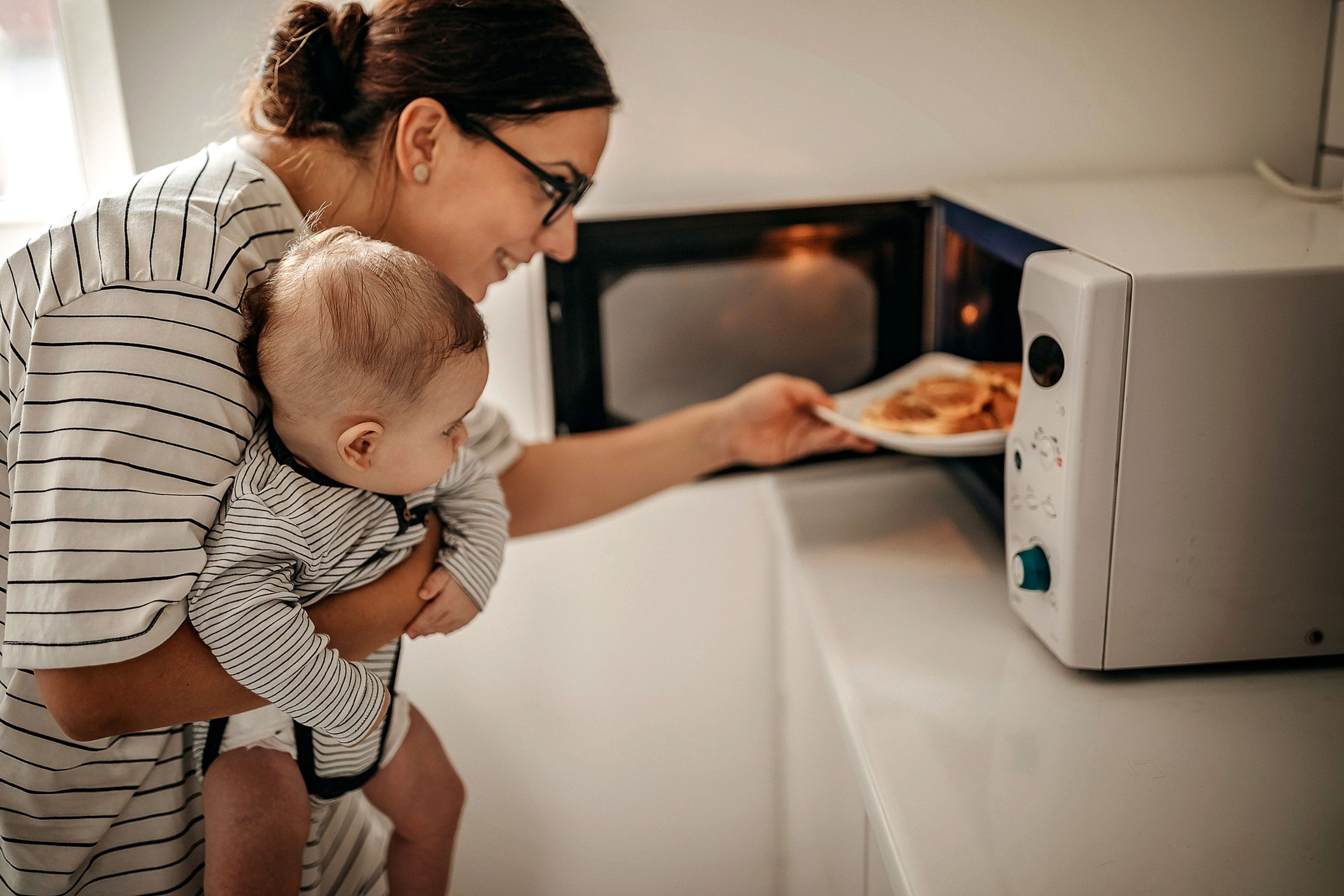 Una mujer mete un plato en el microondas con un bebé en brazos.