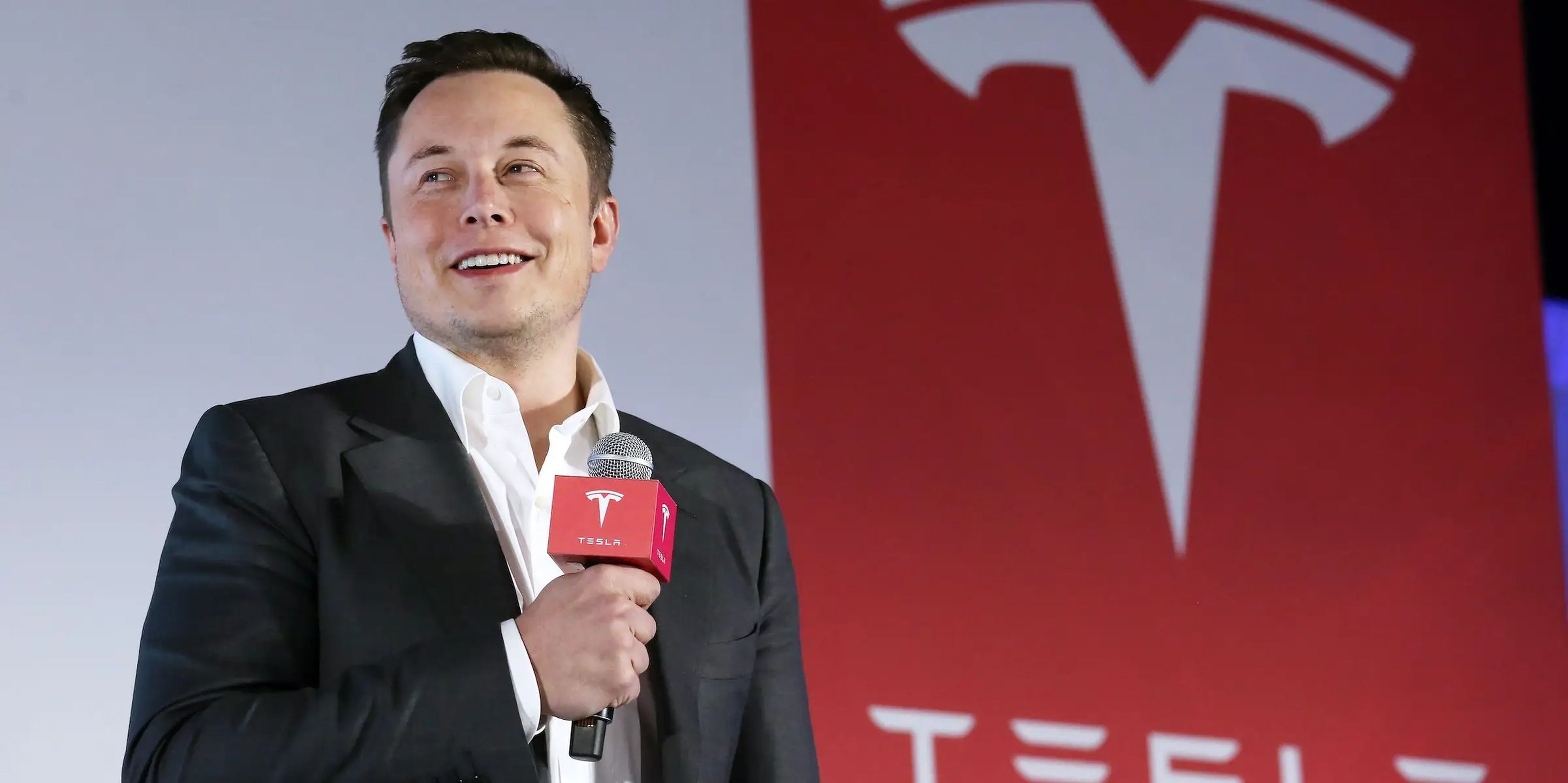 Elon Musk sostiene un micrófono frente a un cartel rojo con el logotipo de Tesla.