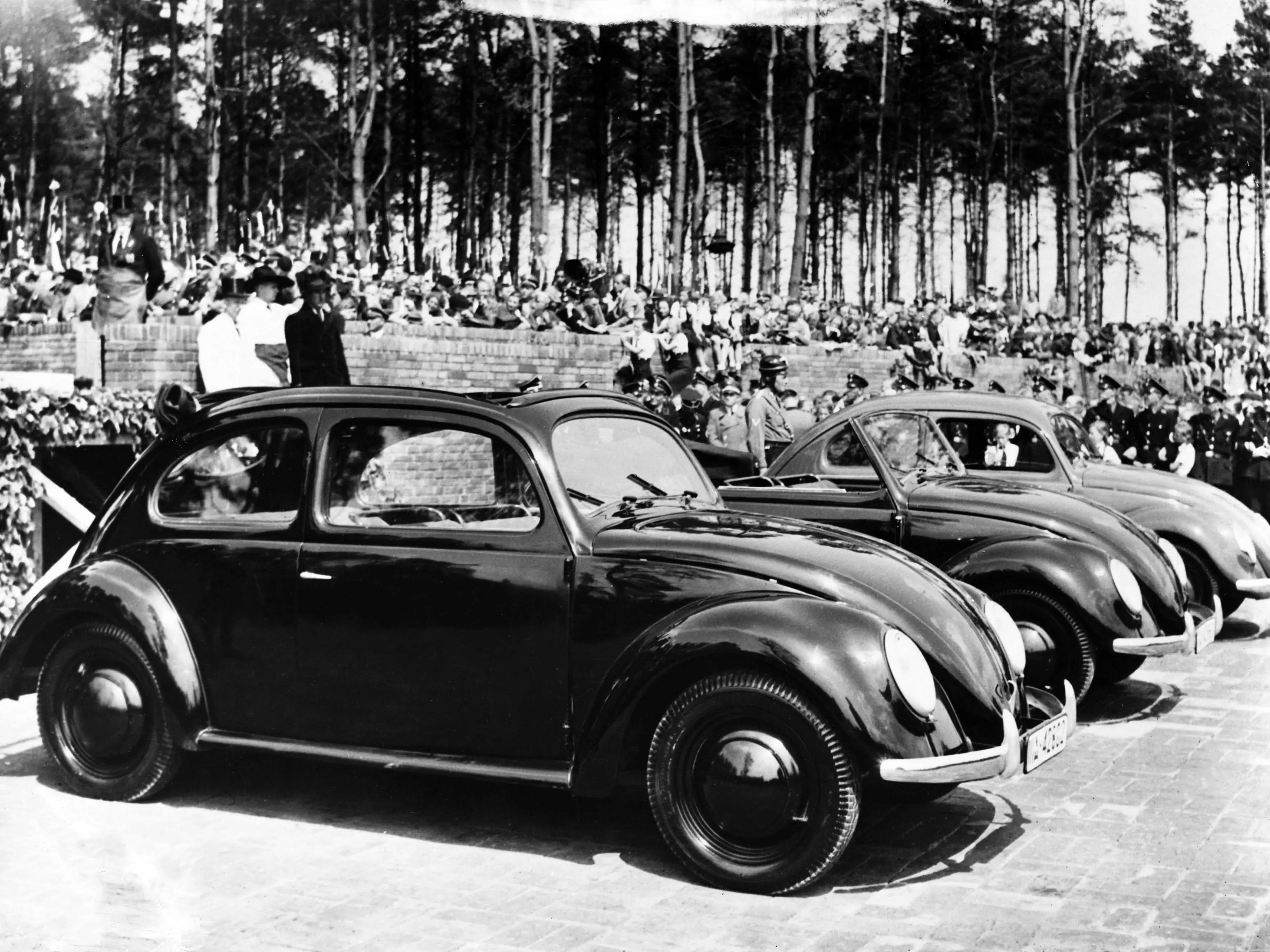 Adolf Hitler está de pie detrás de los coches con un abrigo oscuro, preparándose para inaugurar la nueva fábrica de Volkswagen en Fallersleben.