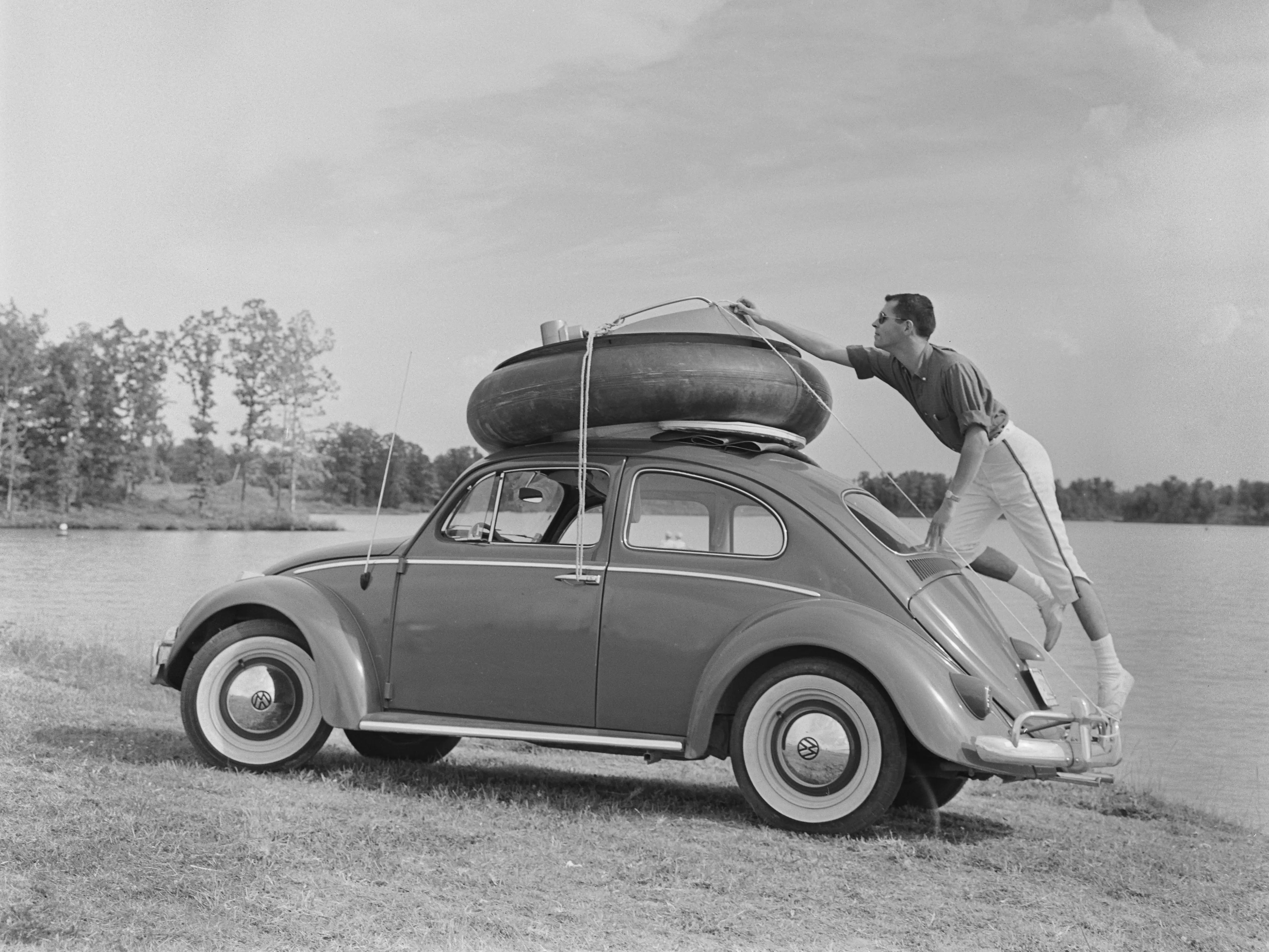 Un hombre se sube al parachoques trasero de un Volkswagen Beetle mientras ajusta las cuerdas que atan una rueda de repuesto y un adaptador de barco al techo del coche. Estados Unidos, alrededor de 1955.