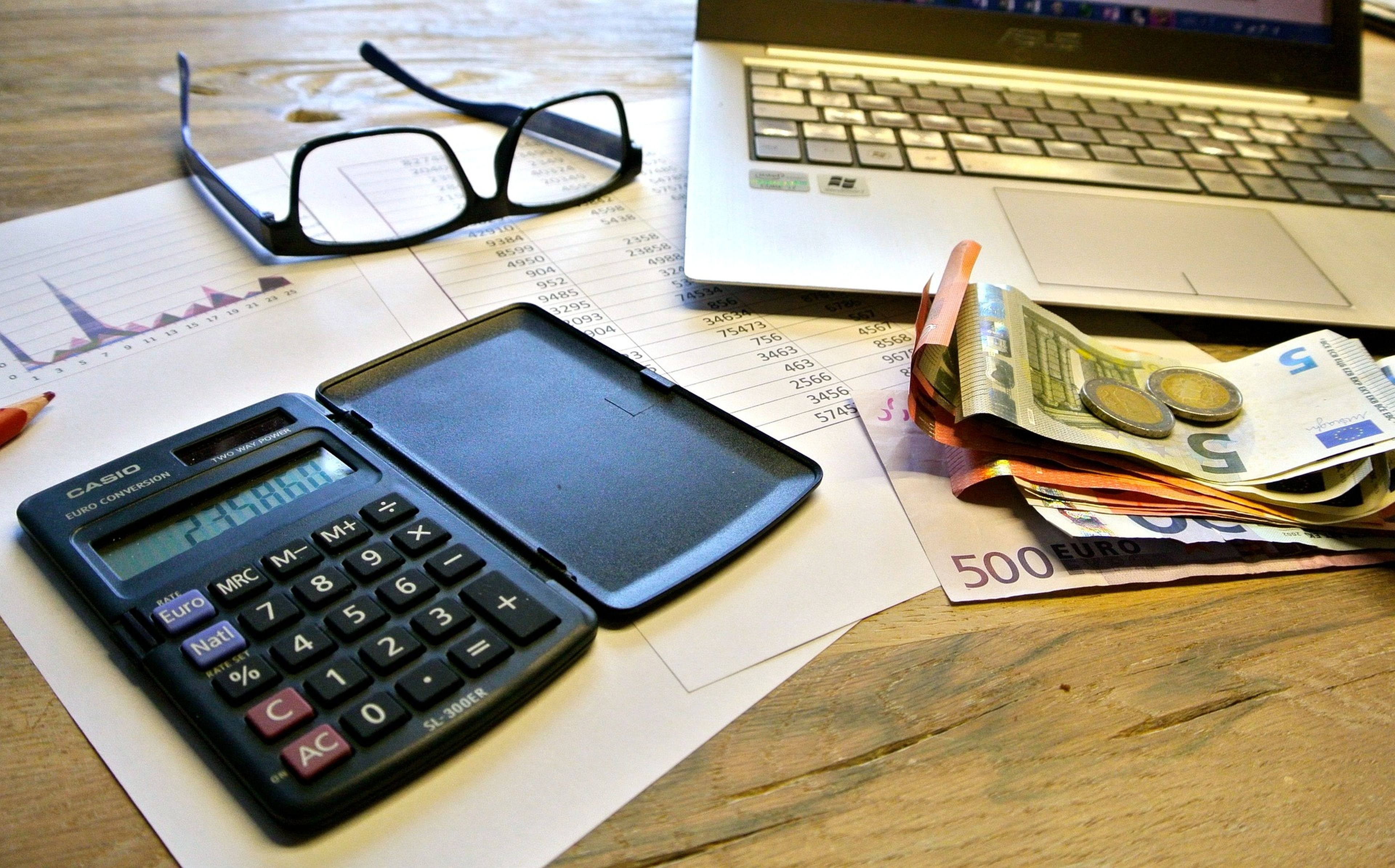 Una mesa con bolígrafos, una calculadora, un ordenador, varios billetes de euro y papeles.