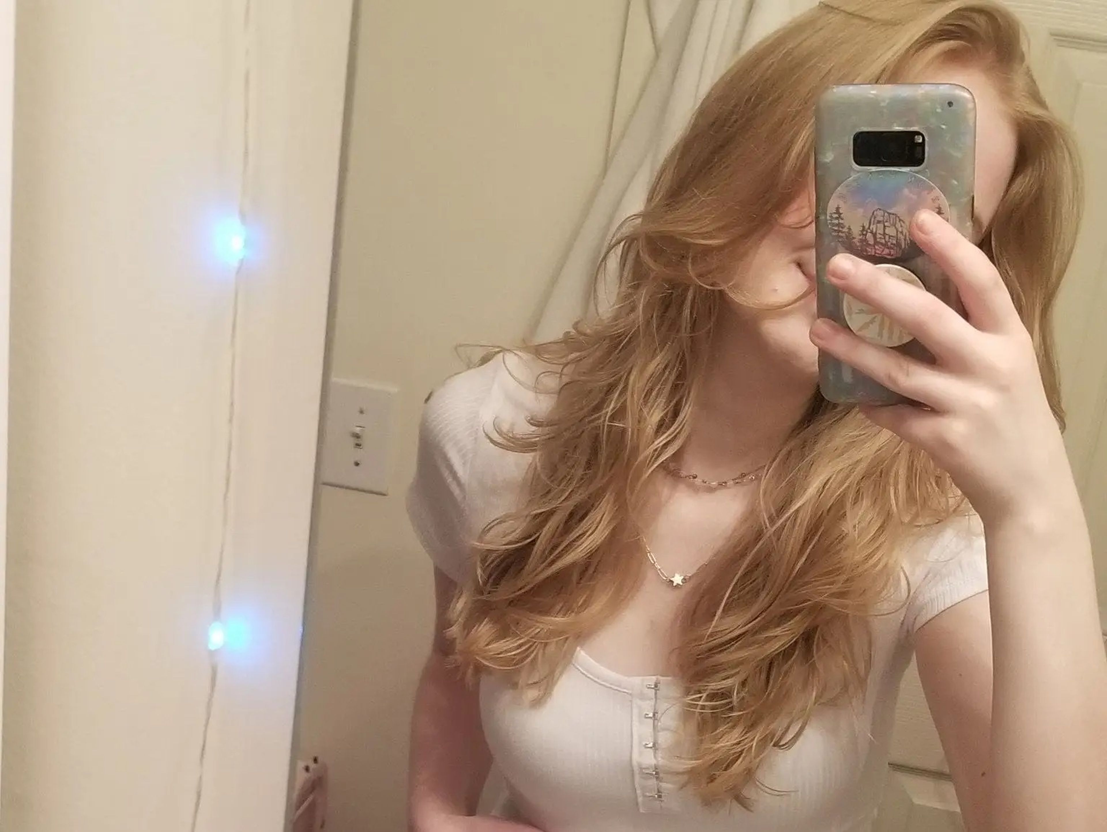 A mirror selfie of Riley, who has strawberry blonde hair and pale skin. Her face is obscured by her phone.