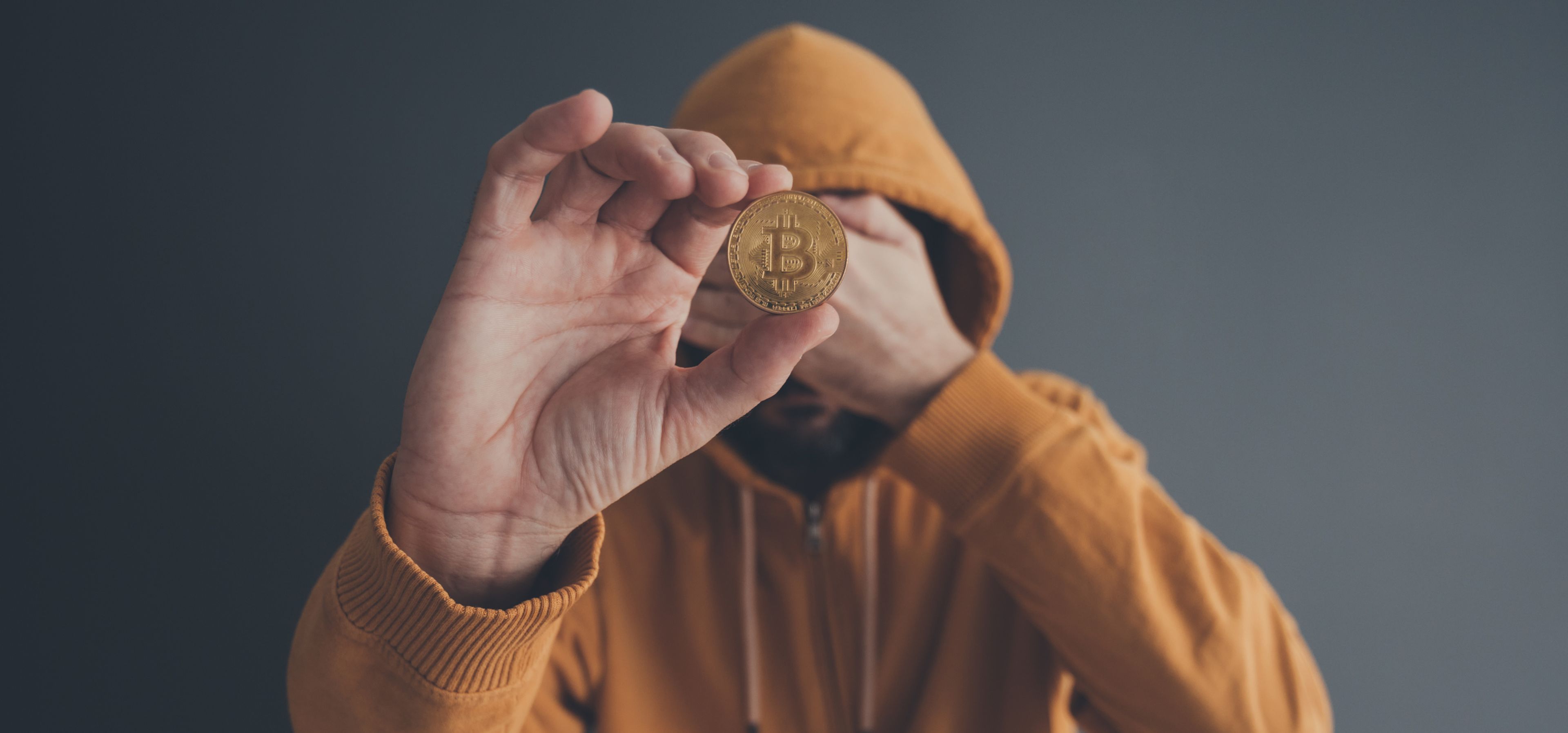 Una persona con cara tapada muestra una moneda de bitcoin. 