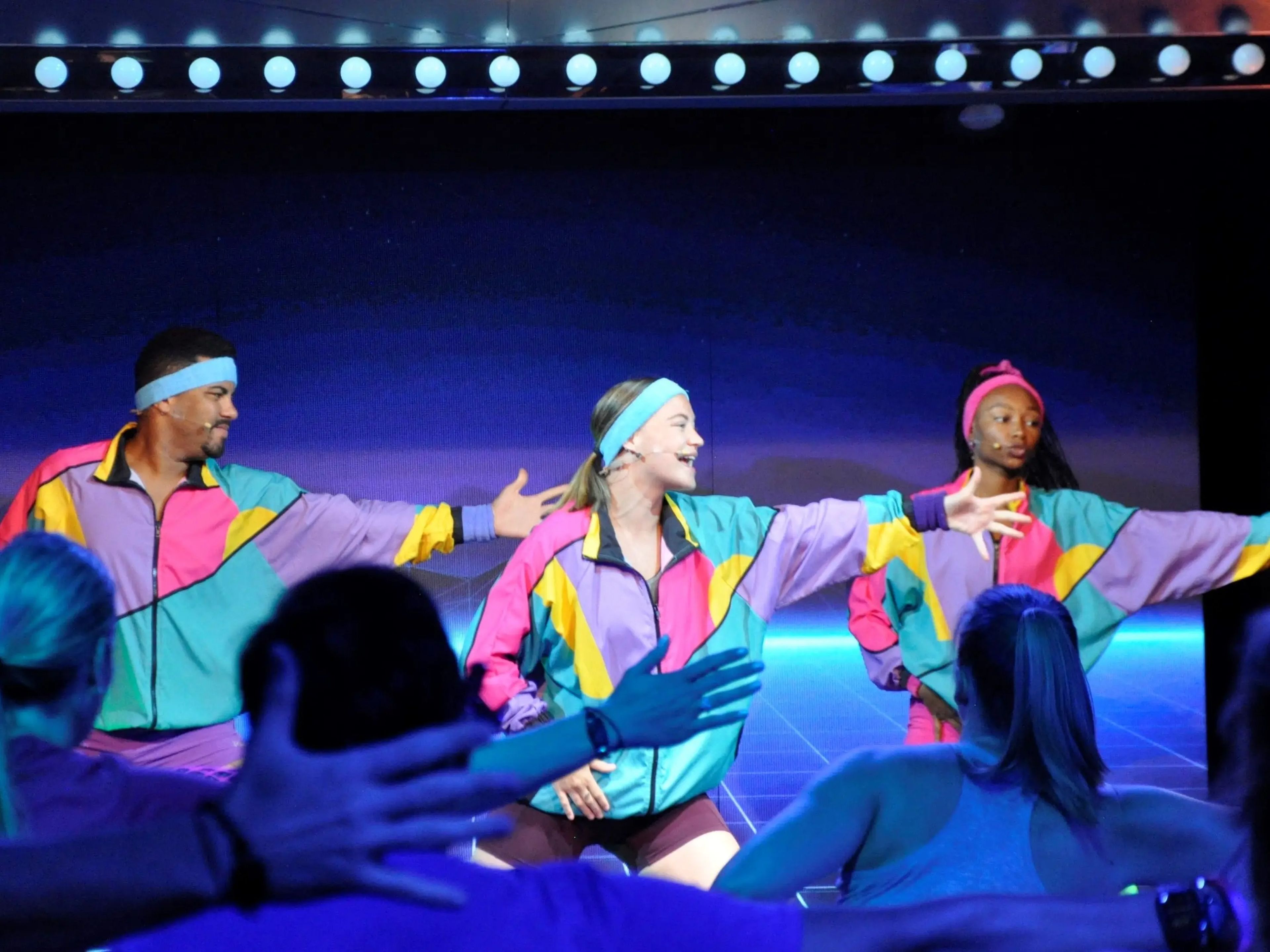 Three people performing in a show wearing neon tracksuits in front of an audience.