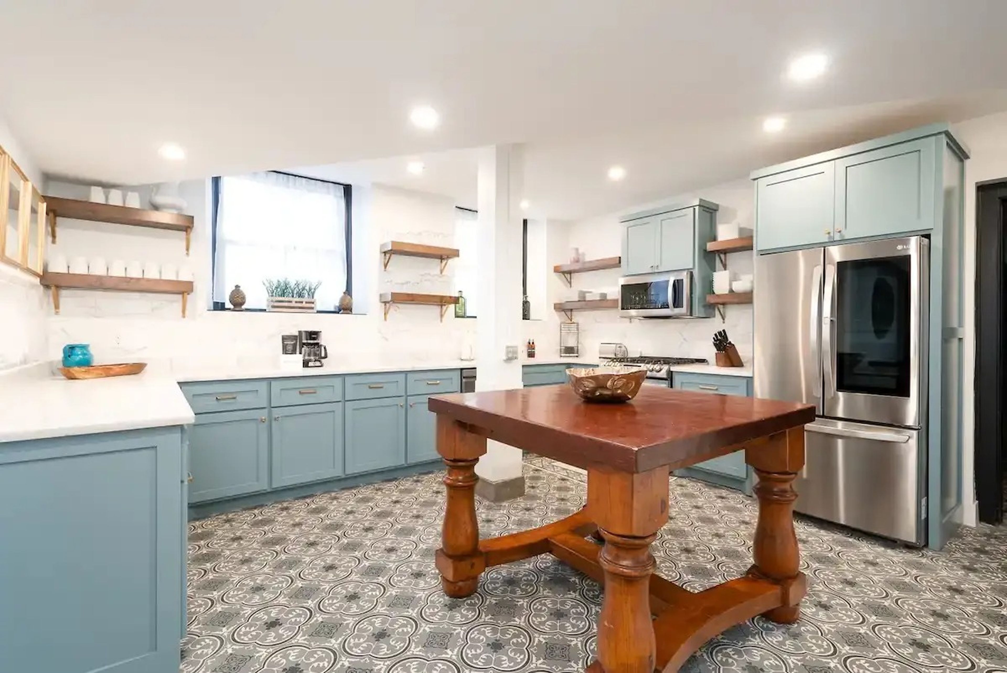 A kitchen with patterned flors, light blue cabinets, white cabinets, and a wood table in the middle.