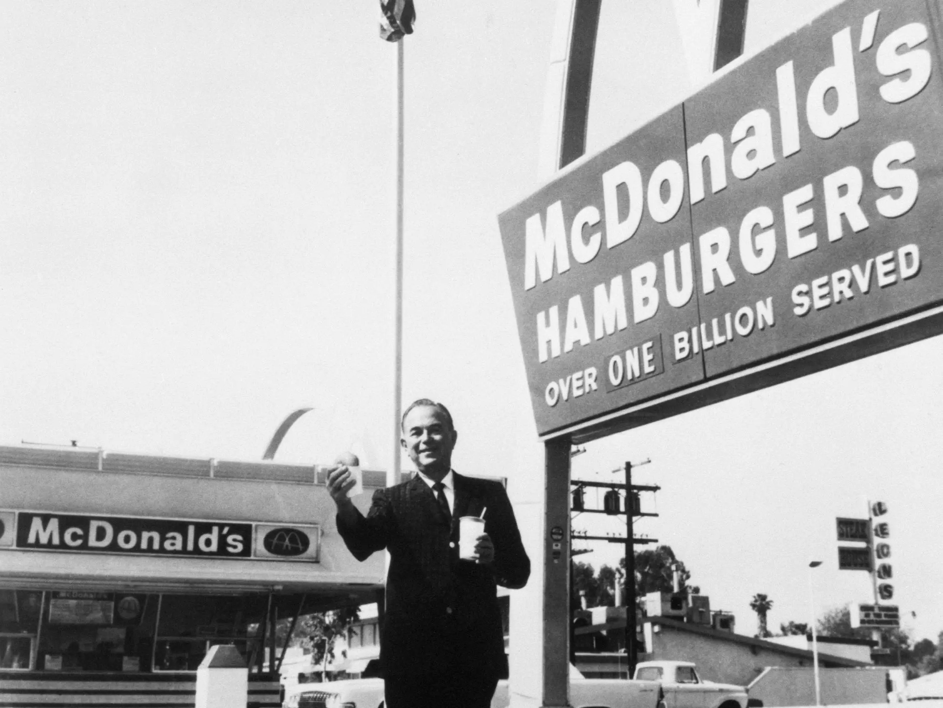 Ray Kroc, fundador y presidente de McDonald's Corporation, delante de una de sus franquicias, con una hamburguesa y una bebida en la mano.