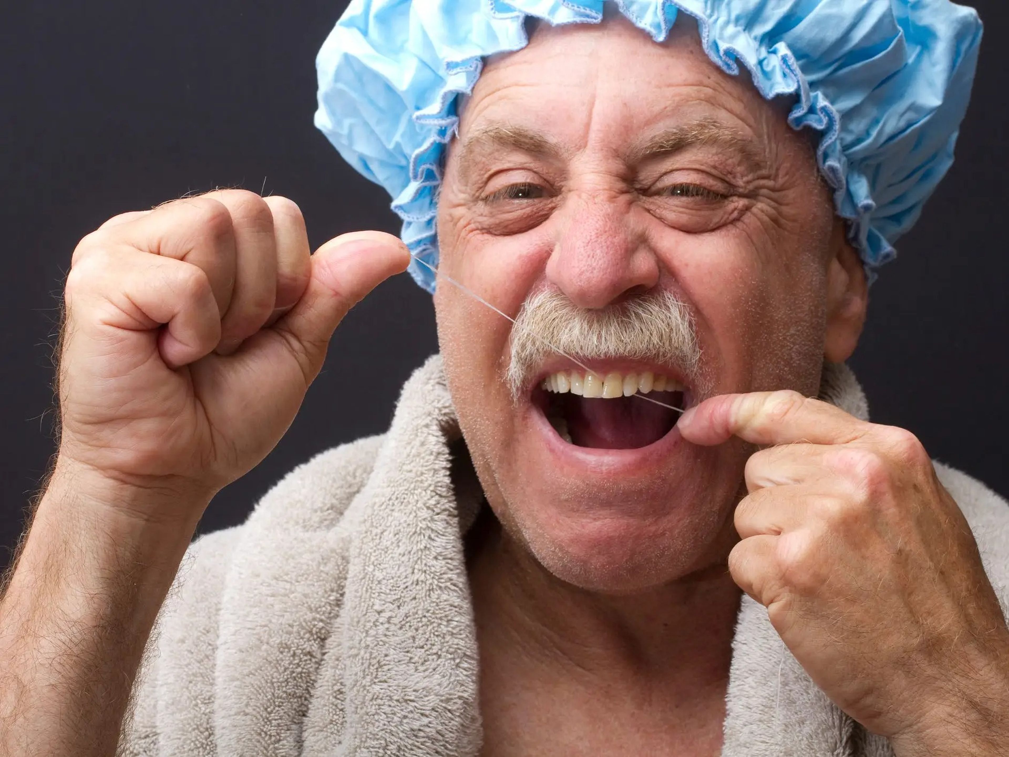 An old man with a thick walrus moustache and a blue shower cap flossing his teeth.