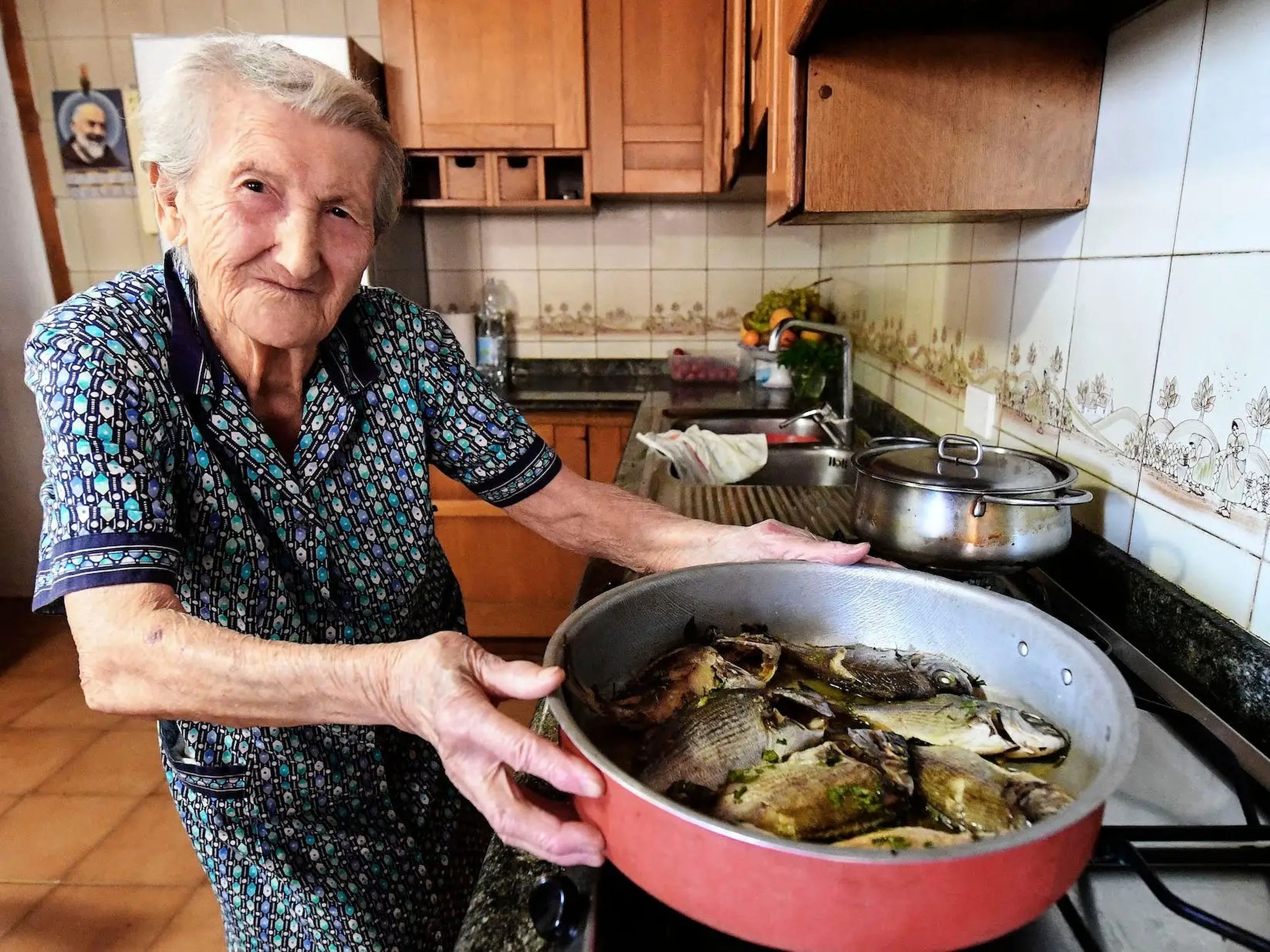 An old woman cooking fish in a large red pan in a small kitchen.