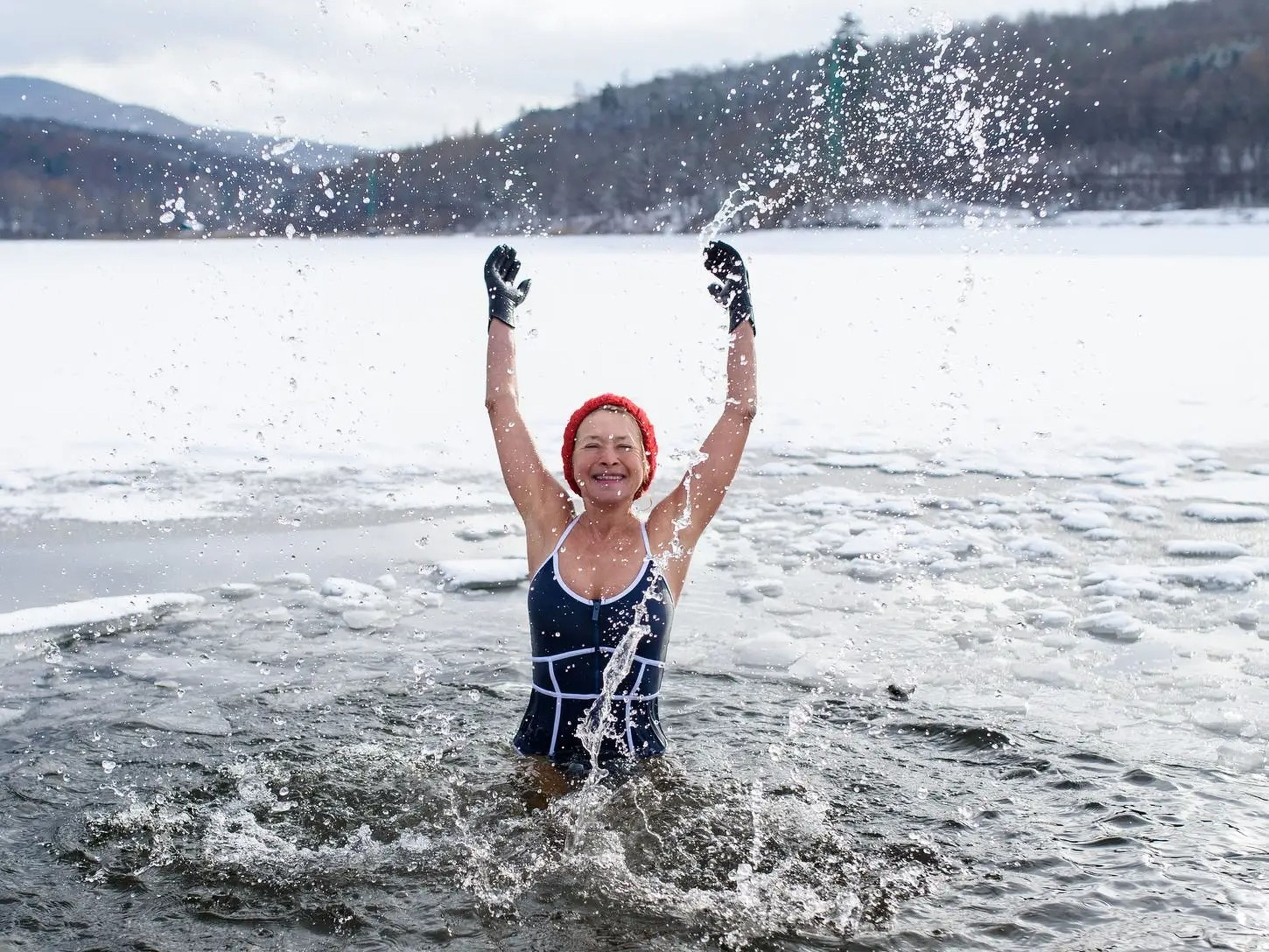 A woman in swimsuit stands in a lake in winter splashing around with a smile on her face.