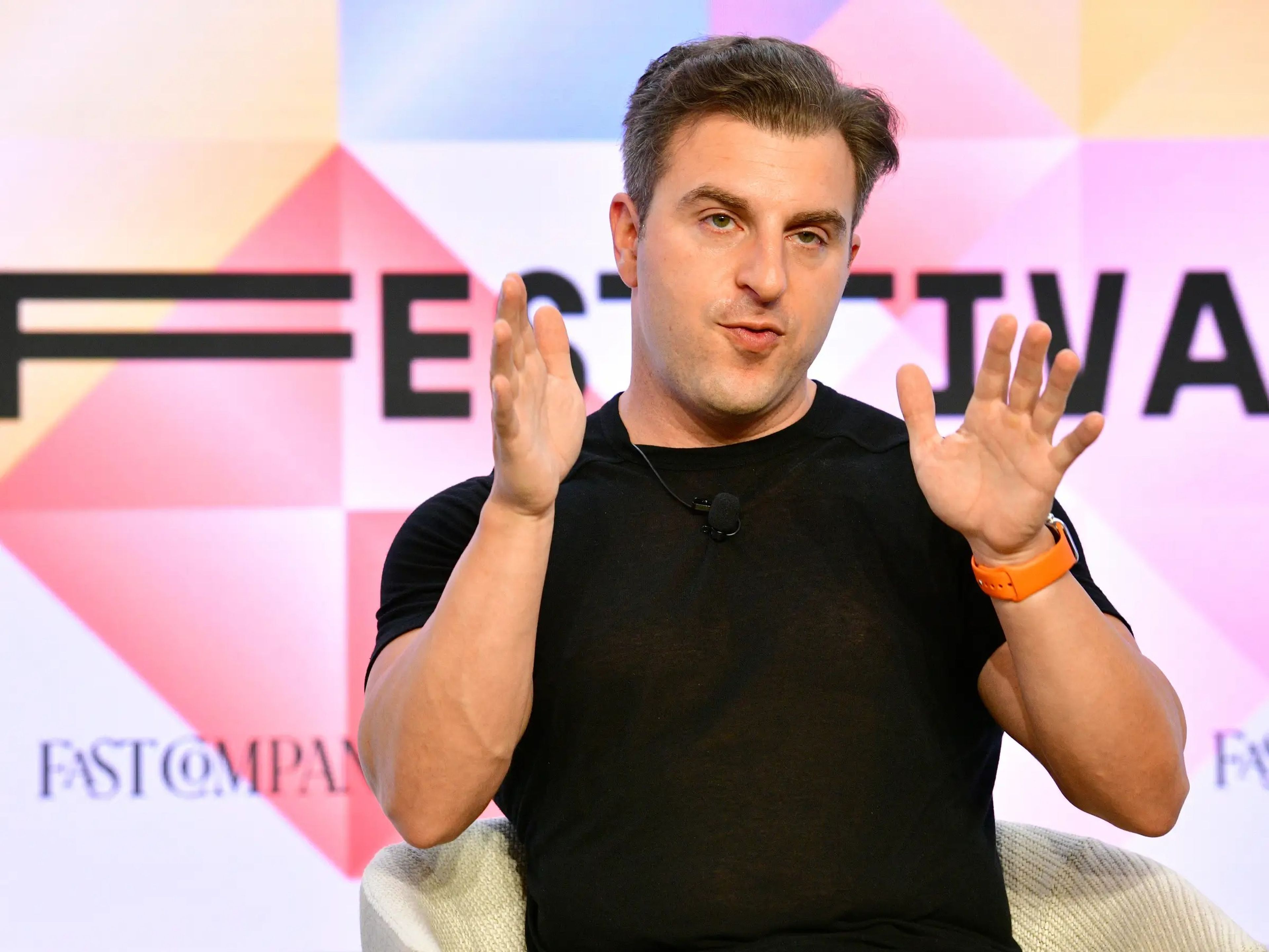 Brian Chesky gesturing with his hands in a black t-shirt in front of a pink, yellow, and blue screen.