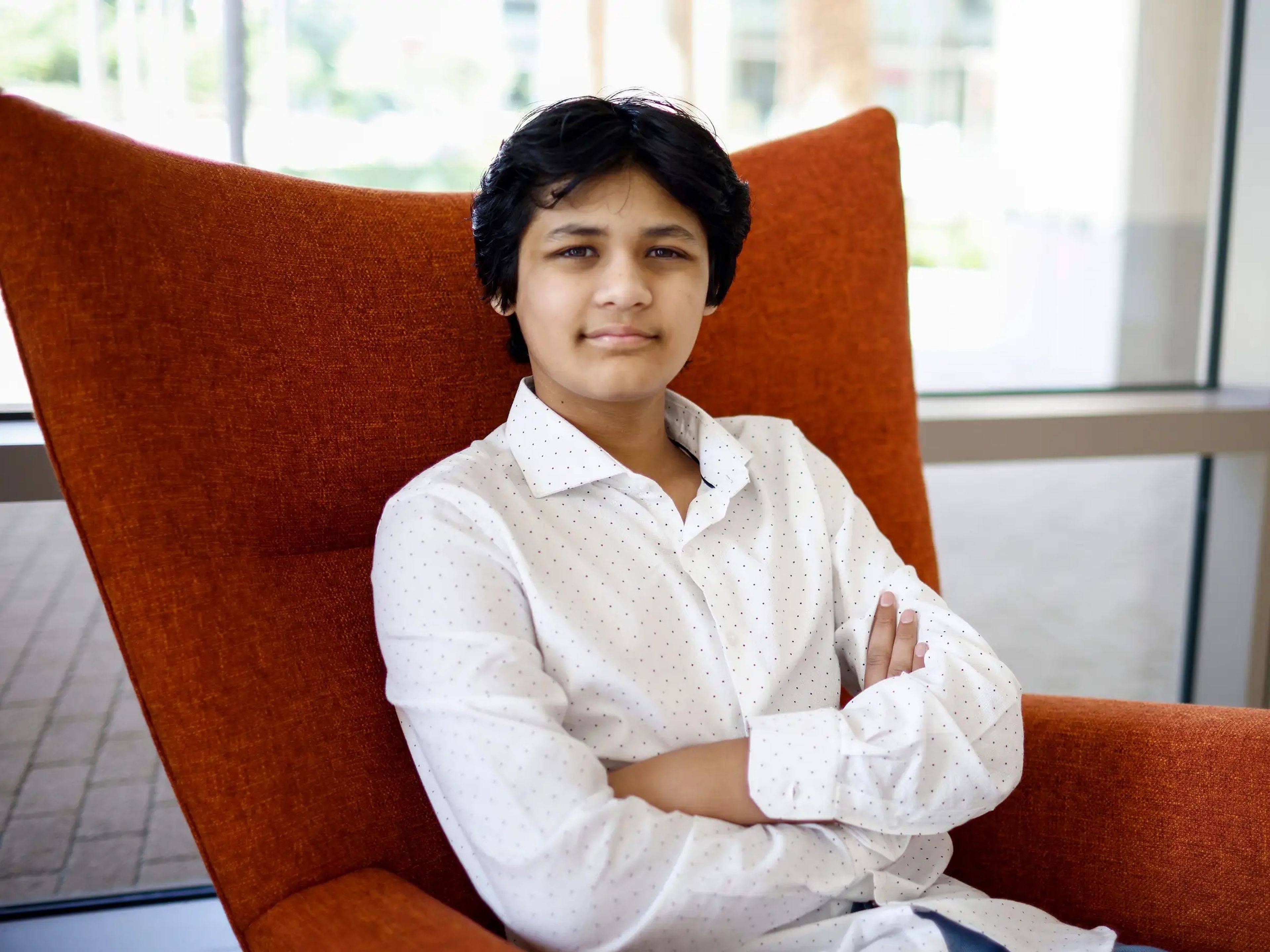 Kairan Quazi wearing a white collared shirt and crossing his arms whilst sat in a red chair