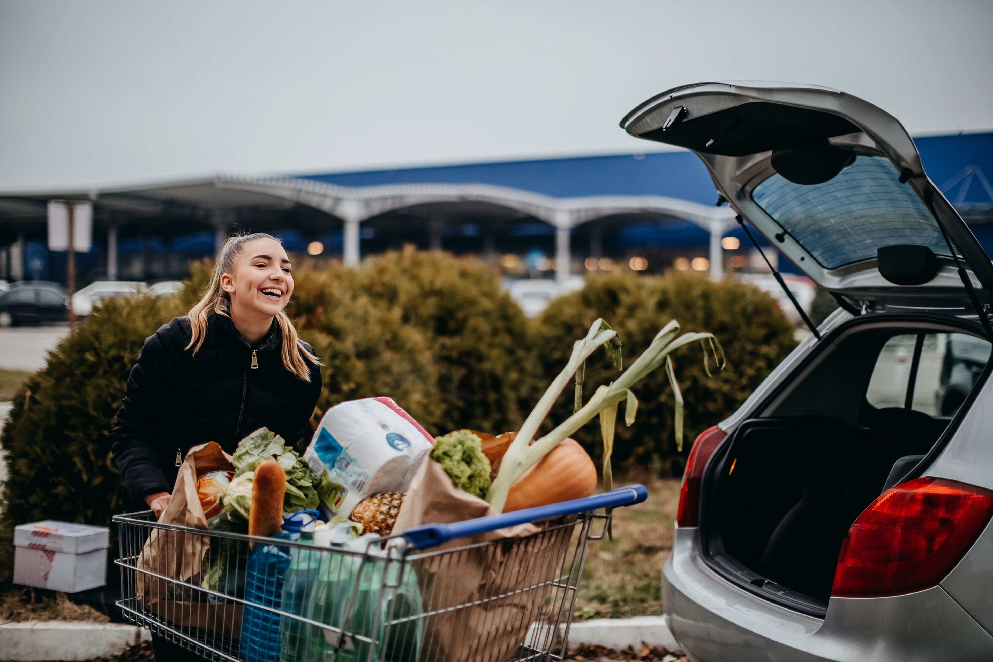 Compra supermercado, chica joven alegre va a su coche con un carro