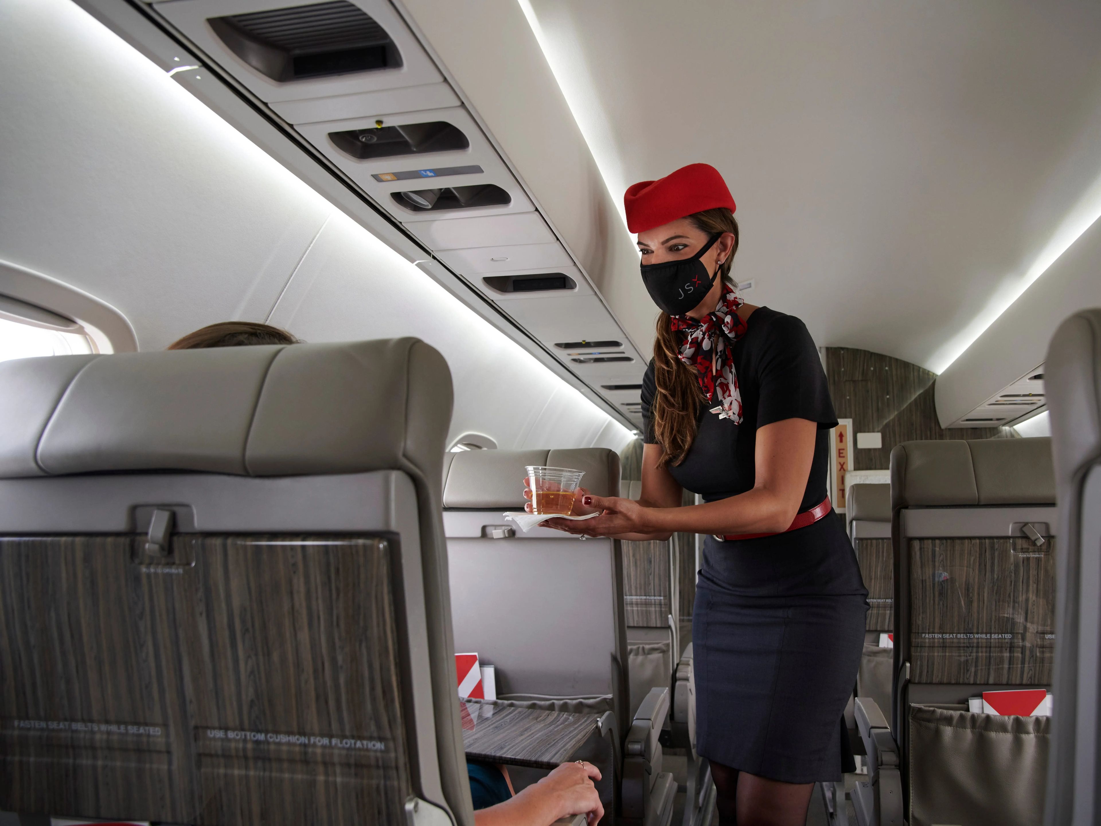 A flight attendant with a face mask serving a drink to a passenger inside a JSX jet.