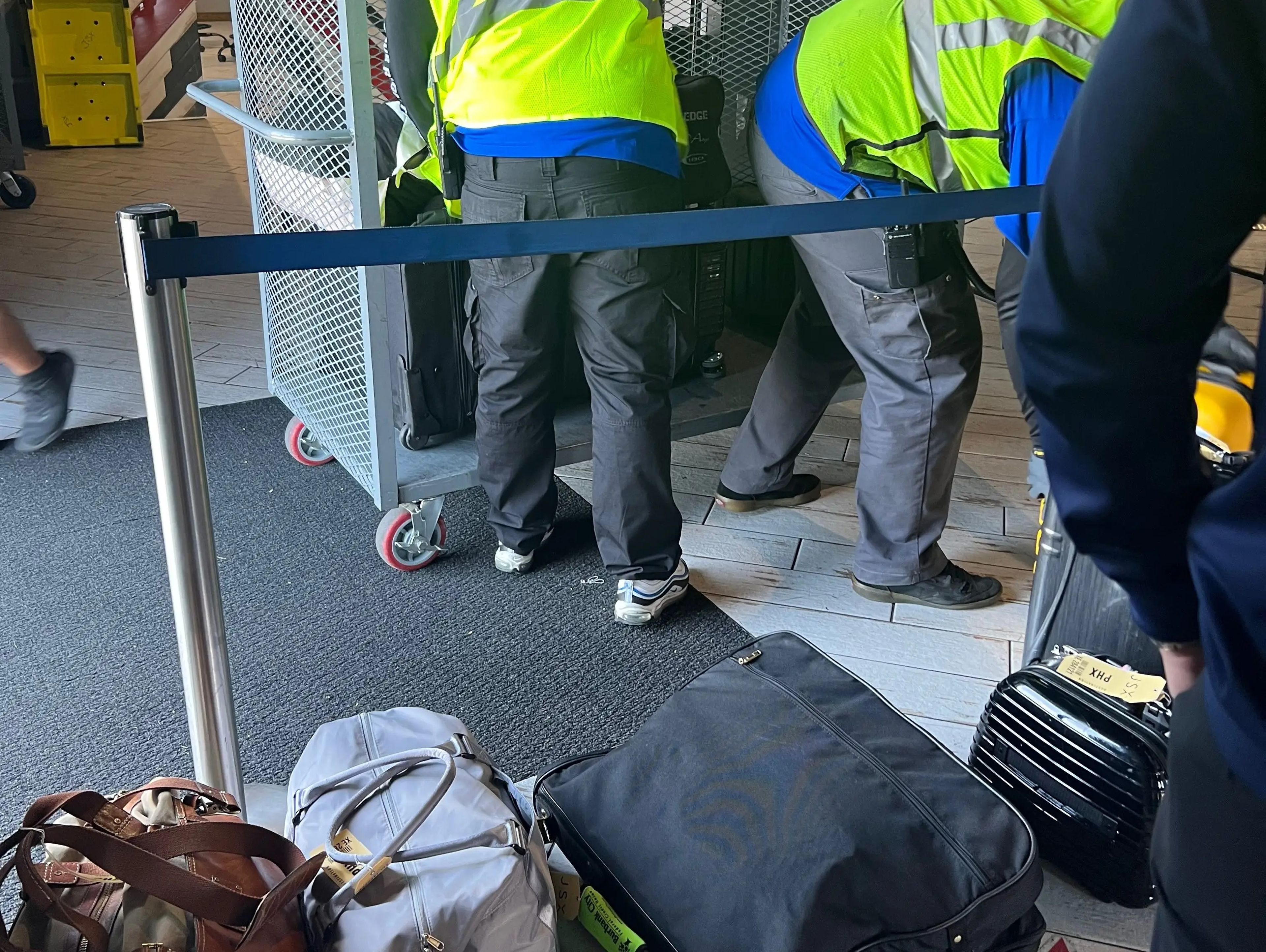 Ground crew in yellow vests standing behind a barrier while unloading luggage from a metal trolley.