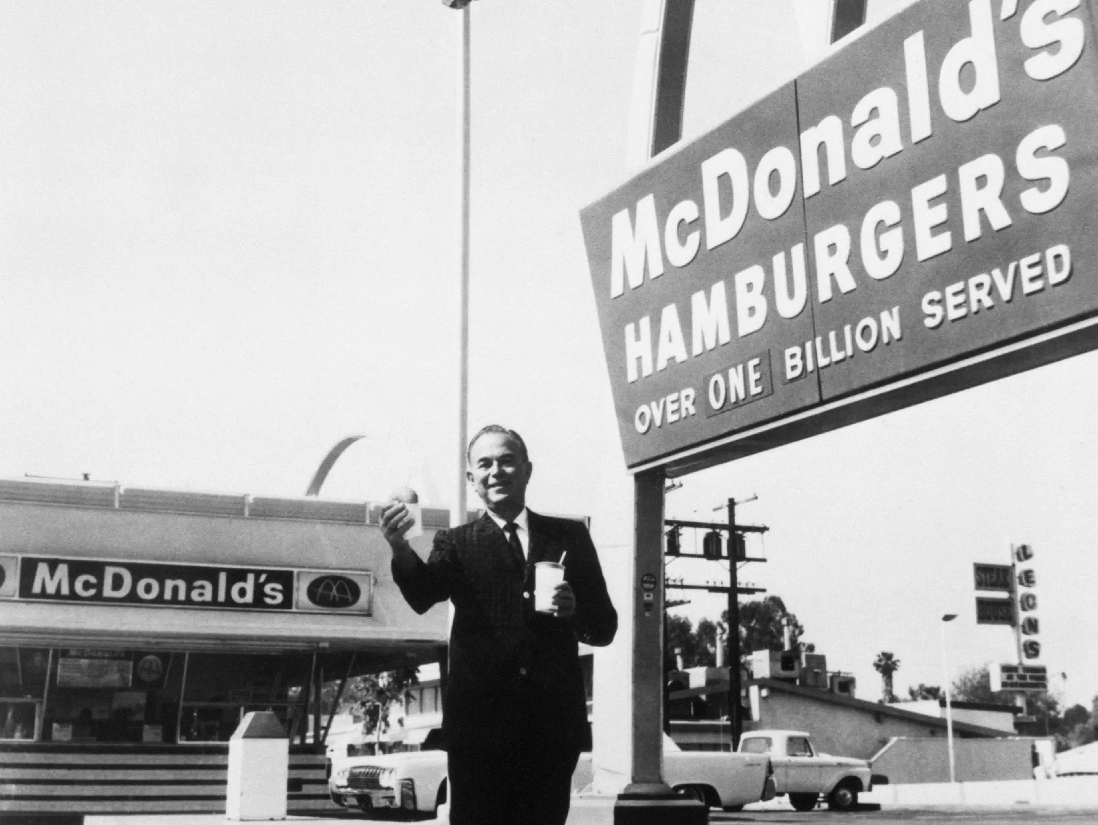 Ray Kroc, fundador y presidente de McDonald's Corporation, delante de una de sus franquicias, con una hamburguesa y una bebida en la mano.