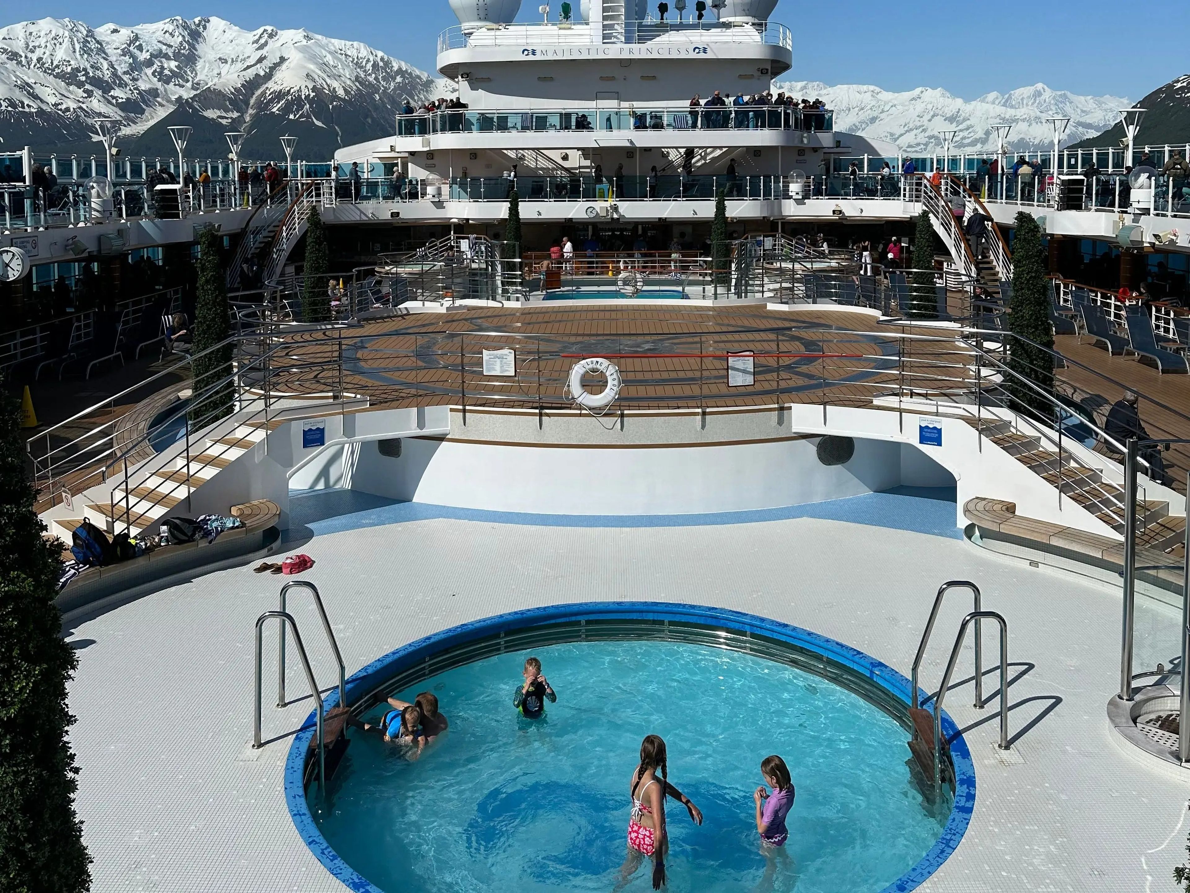 cruise deck with round pool with children on it with snowy mountains in the background