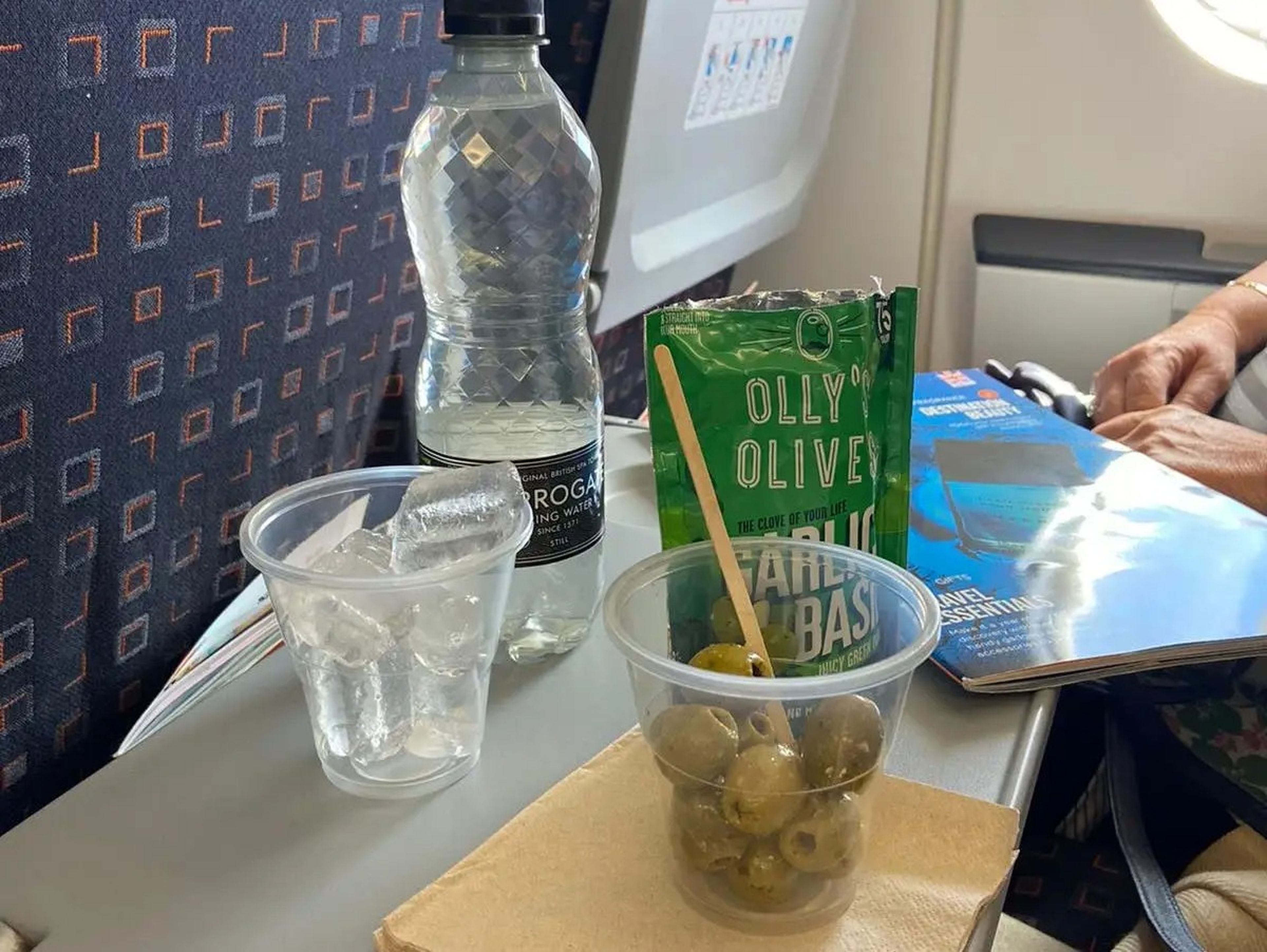 Cup of olives next to empty package of olives and a plastic bottle of beverage on a plane tray