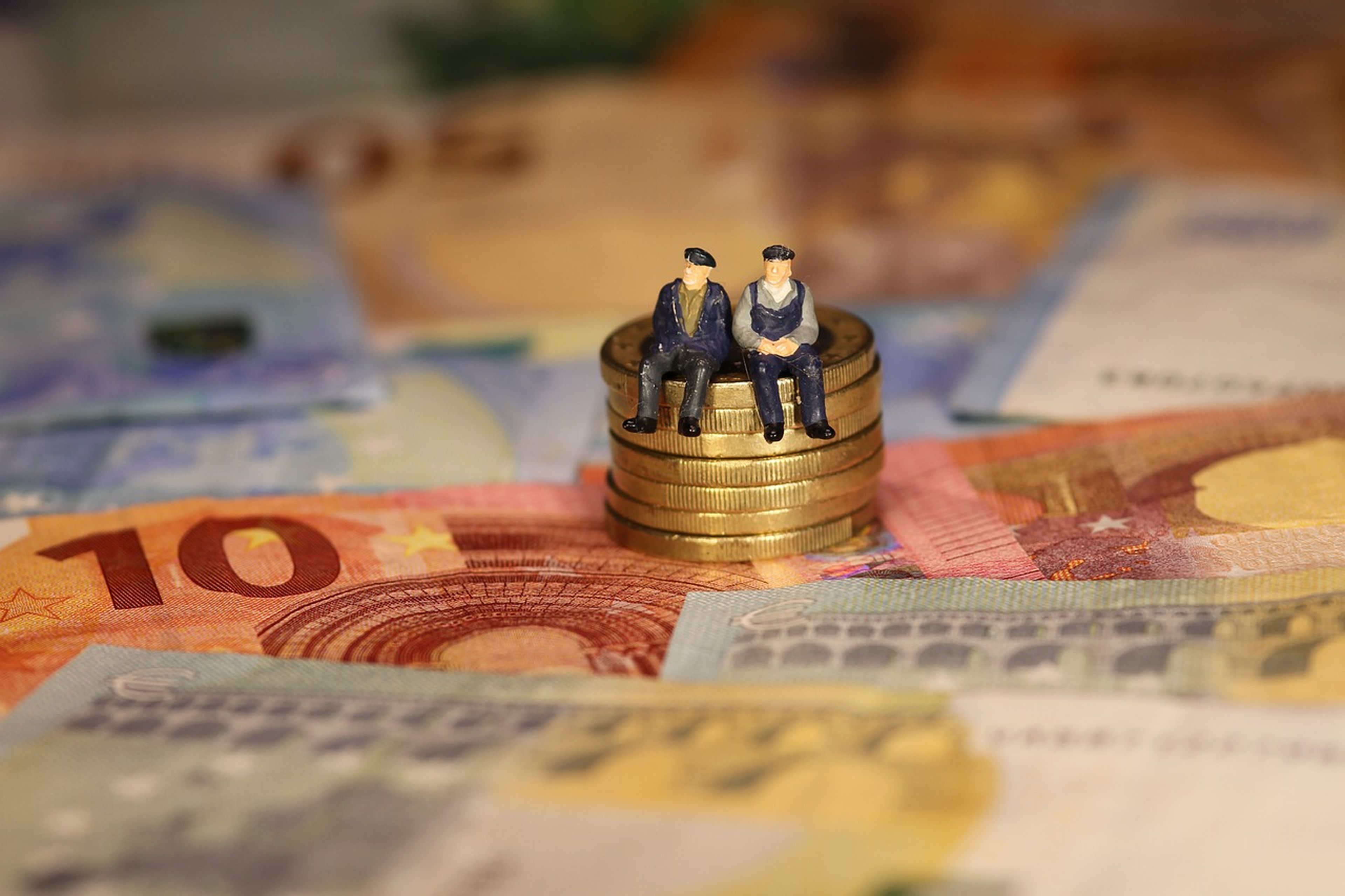 Dos muñecos de personas mayores sentados encima de unas monedas y unos billetes de euros.