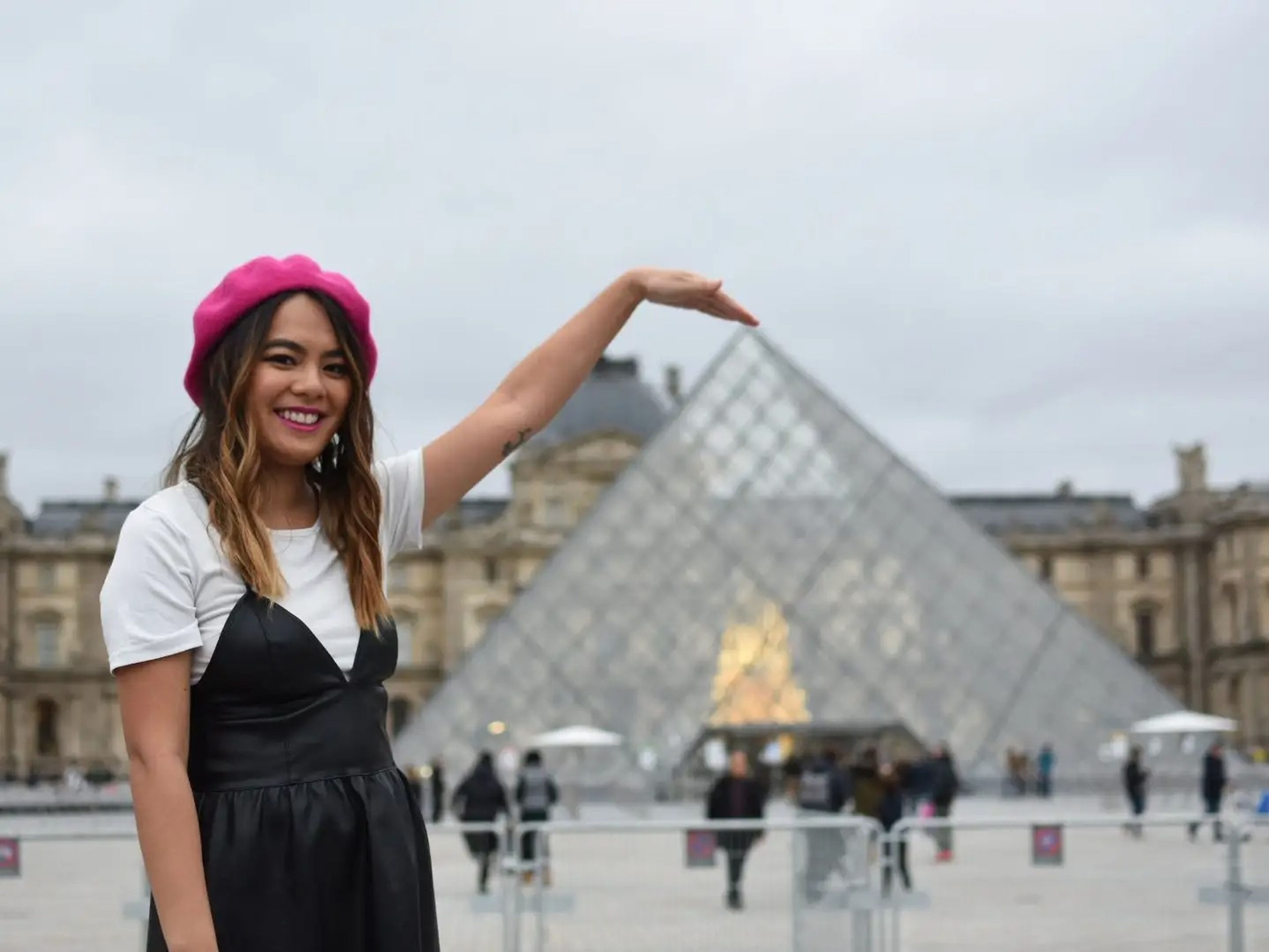 The author wearing a pink beret and a black dress over a white shirt. She is posing in front of the Louvre.
