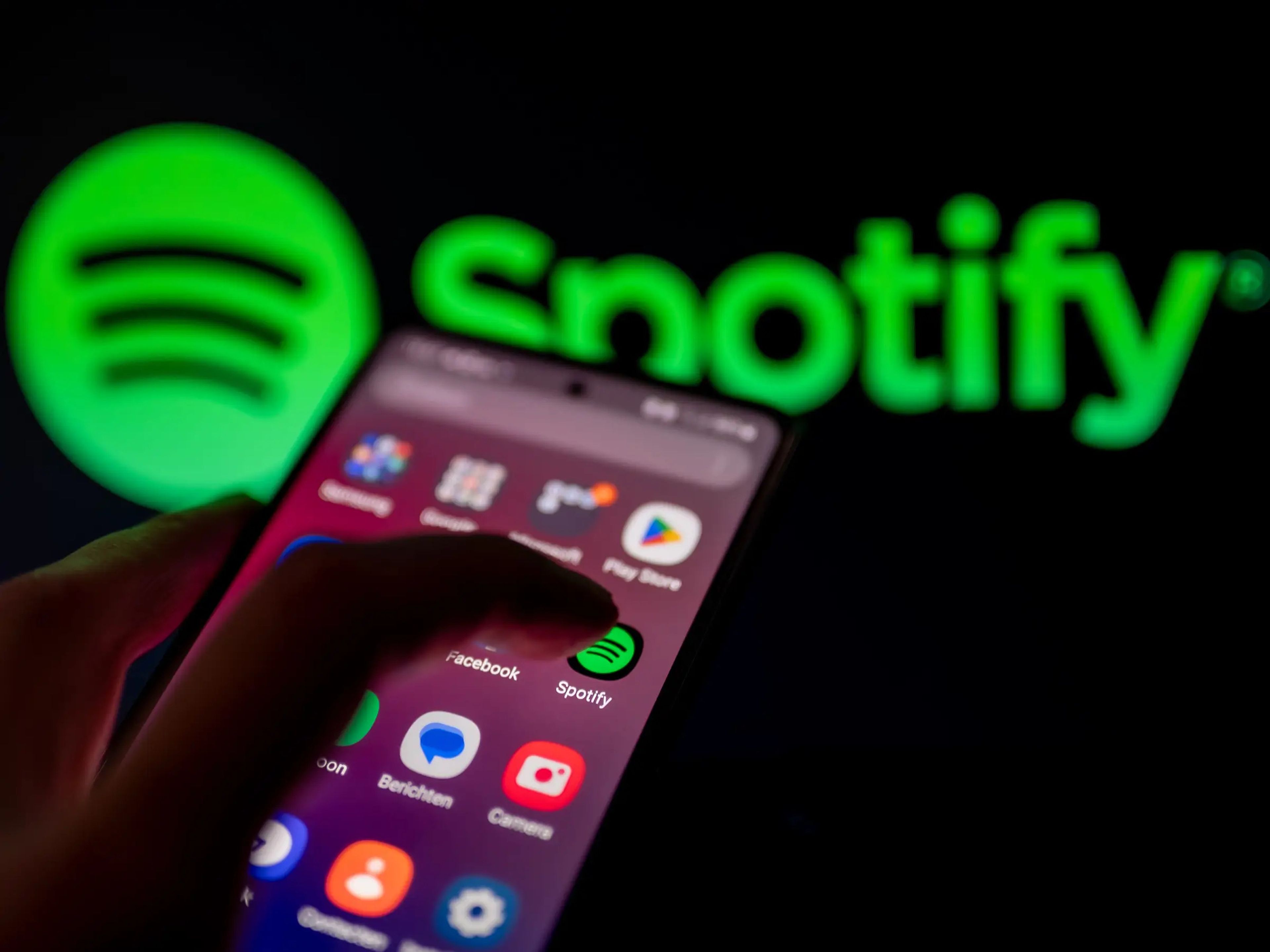 Finger taps the Spotify app on a phone with the Spotify logo blown up in the background.