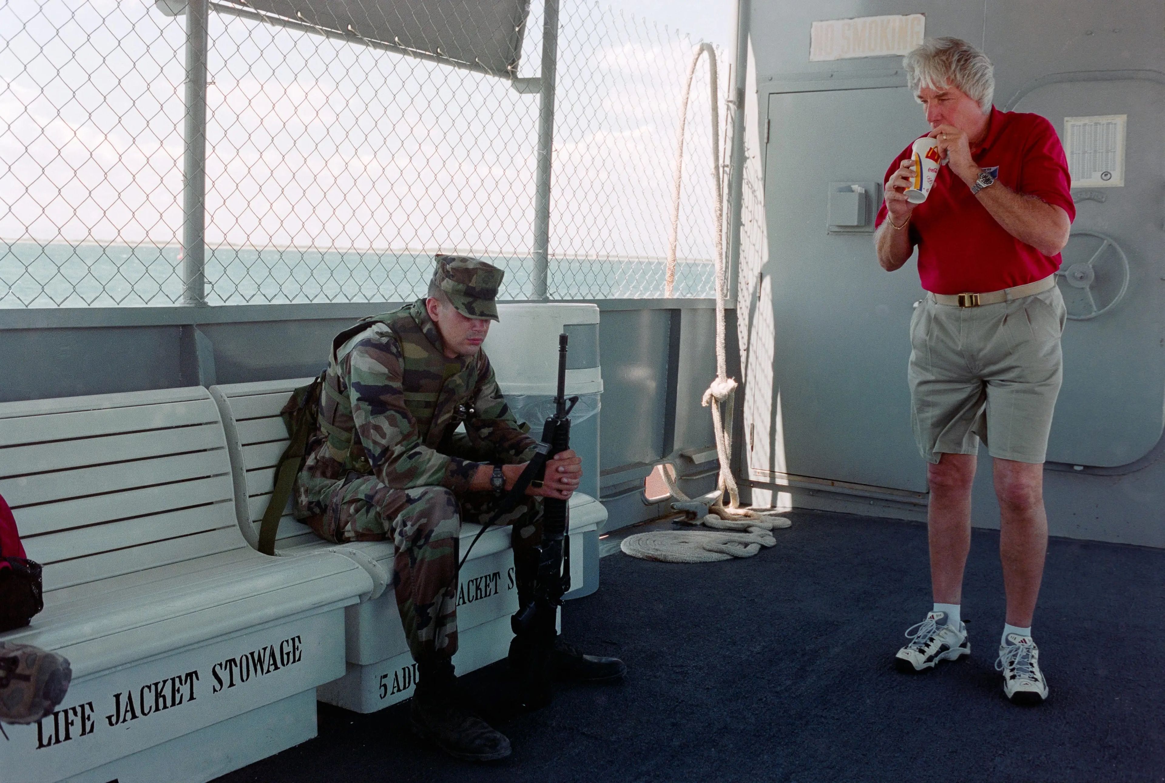 Un soldado de la guarnición de la Joint Task Force 160 vigilando a los prisioneros acompañado de un trabajador civil. Esta imagen fue tomada a bordo del transbordador que conecta los lados este y oeste de la base militar estadounidense.