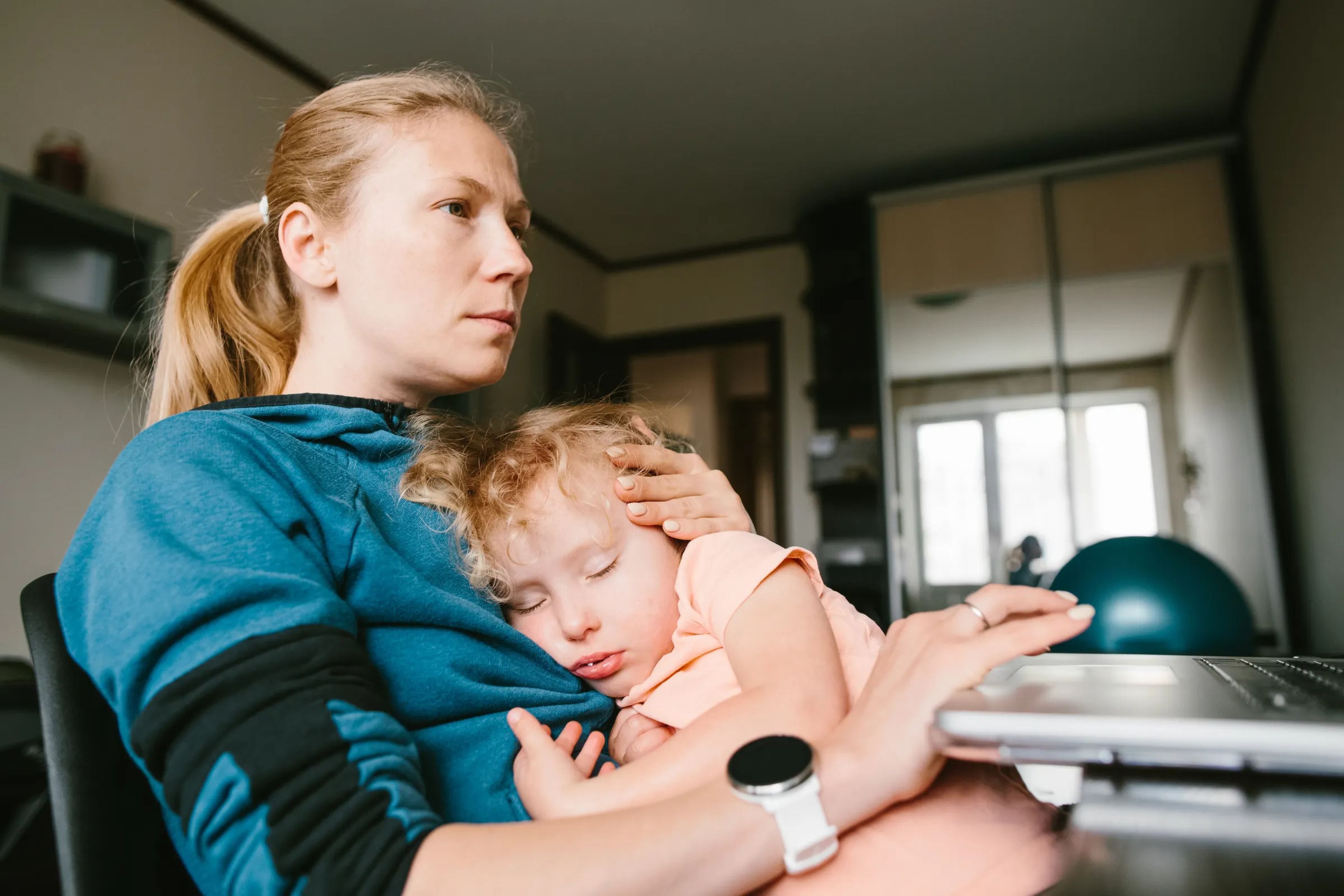 A woman with blonde hair who is wearing a blue sweatshirt looks at a computer while a child in a salmon t-shirt sleeps in her lap. 