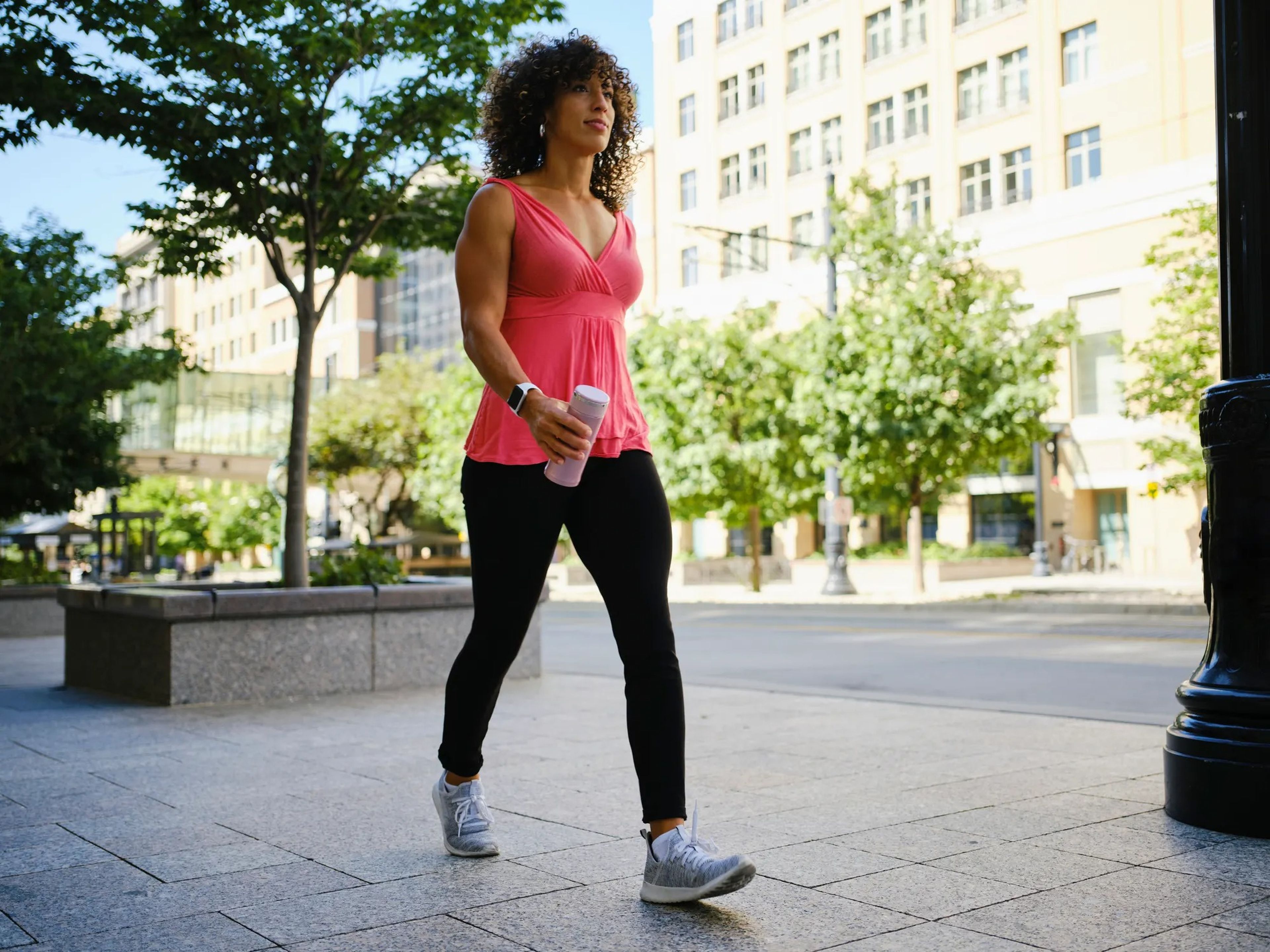 a woman walking a walk in an urban park area, wearing a pink tank top and black leggings and carrying a thermos