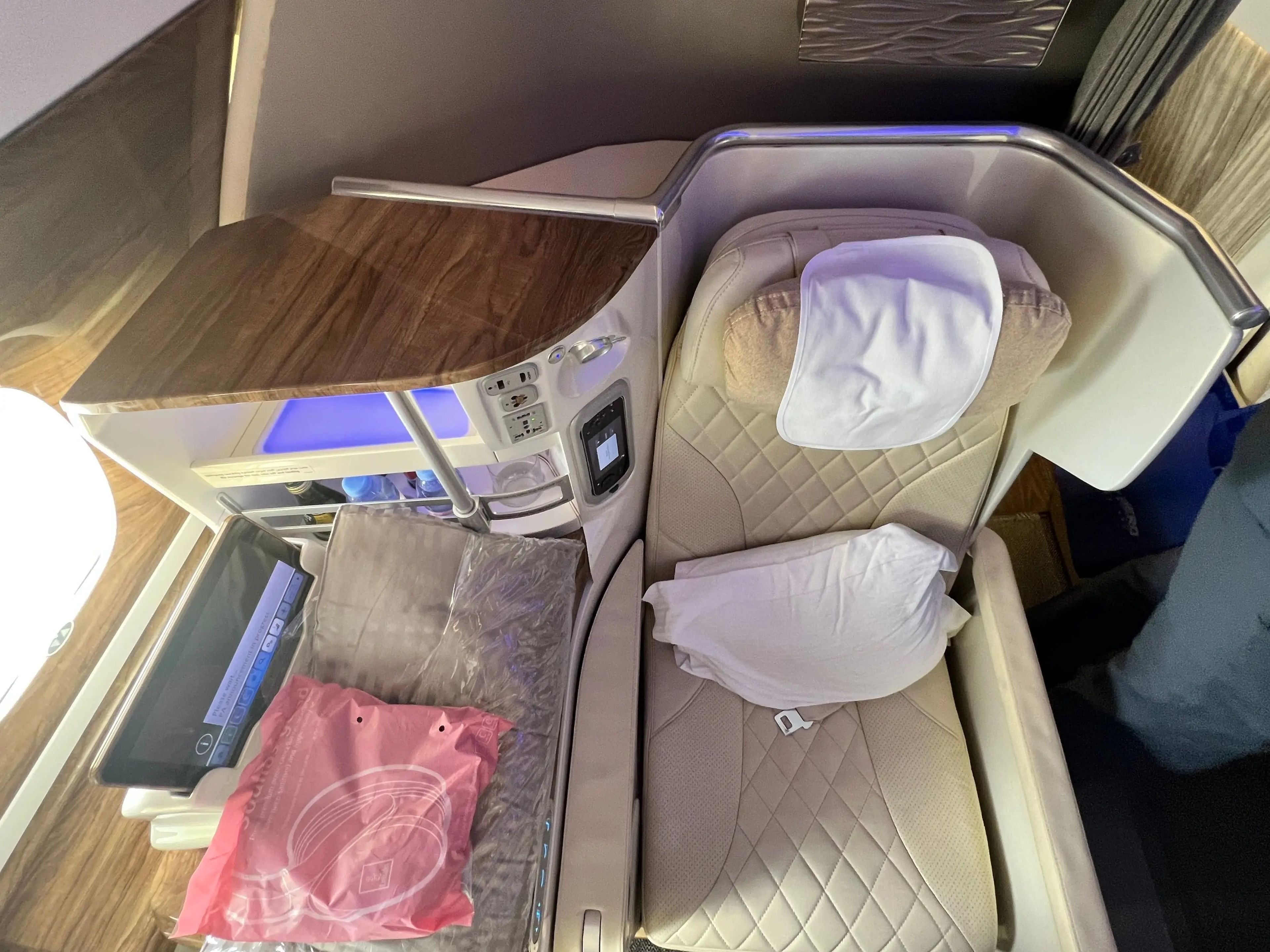 A business class seat in beige leather with white cushion, amenities and drinks holder on the side, onboard Emirates A380