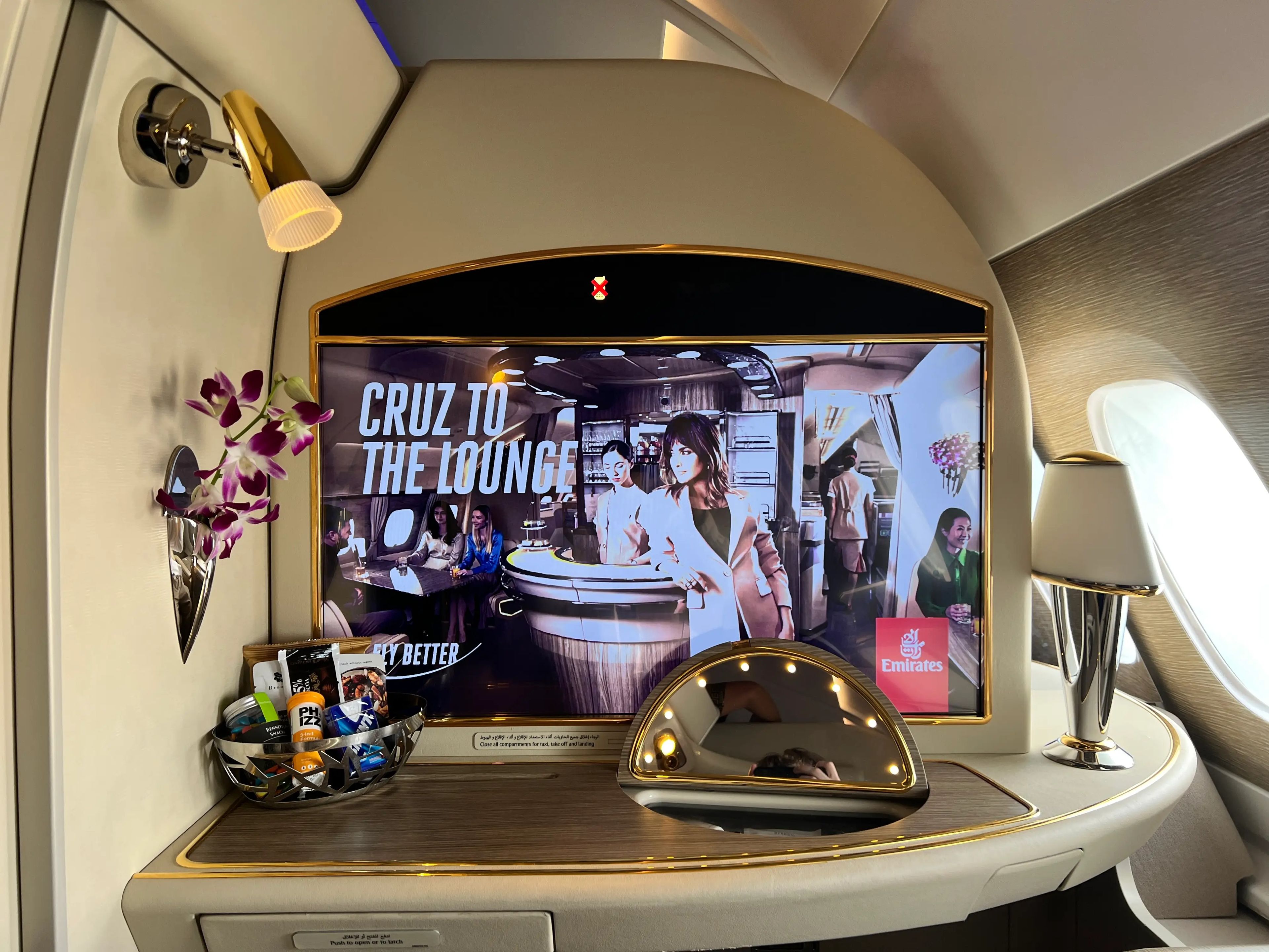 A close up of the 32-inch screen in an Emirates Airbus A380 with two lamps, a pop-up mirror, and basket of snacks
