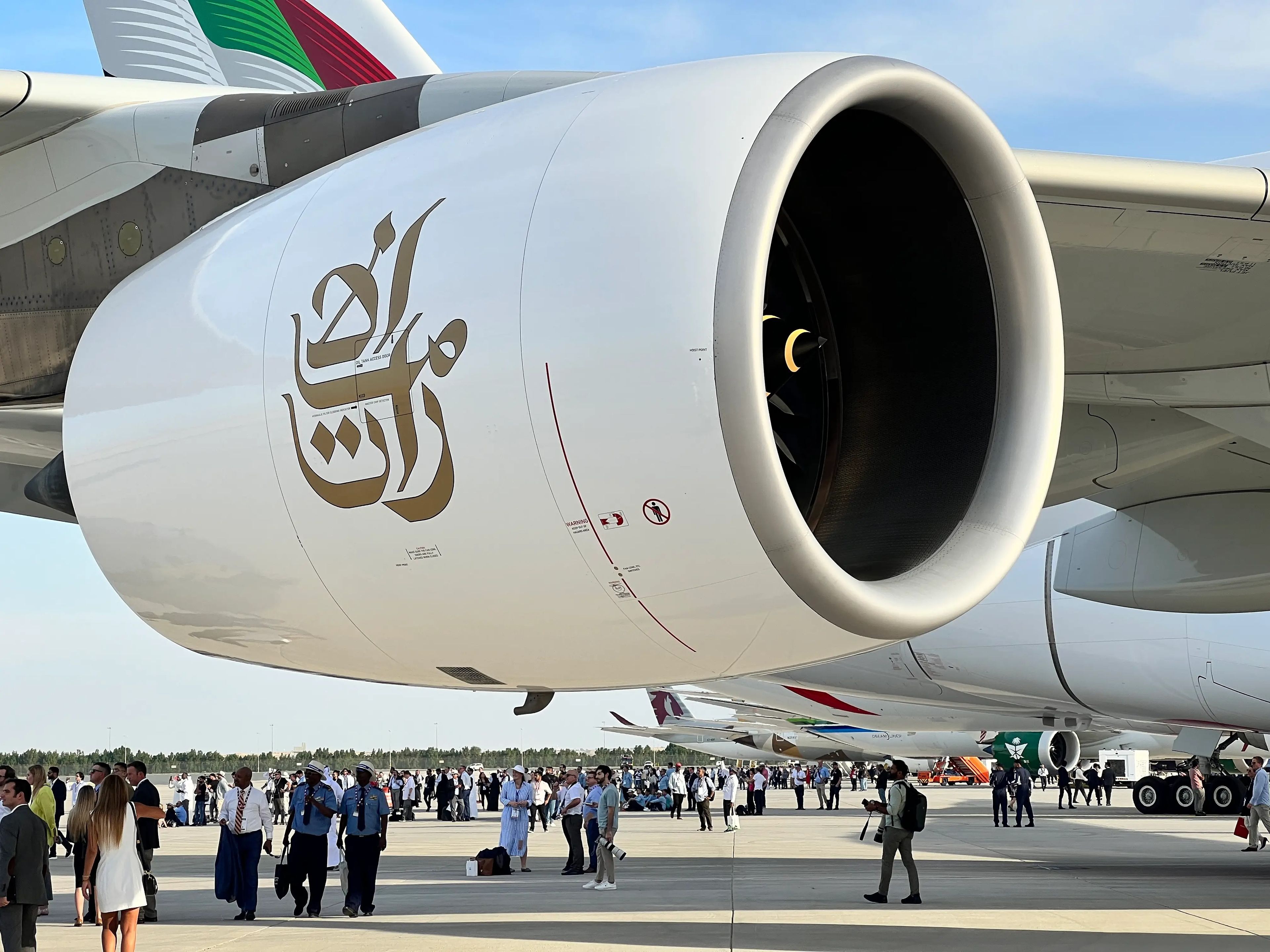 A close-up of an engine on an Emirates Airbus A380-800 at the Dubai Airshow, with people walking underneath and a crowd in the background