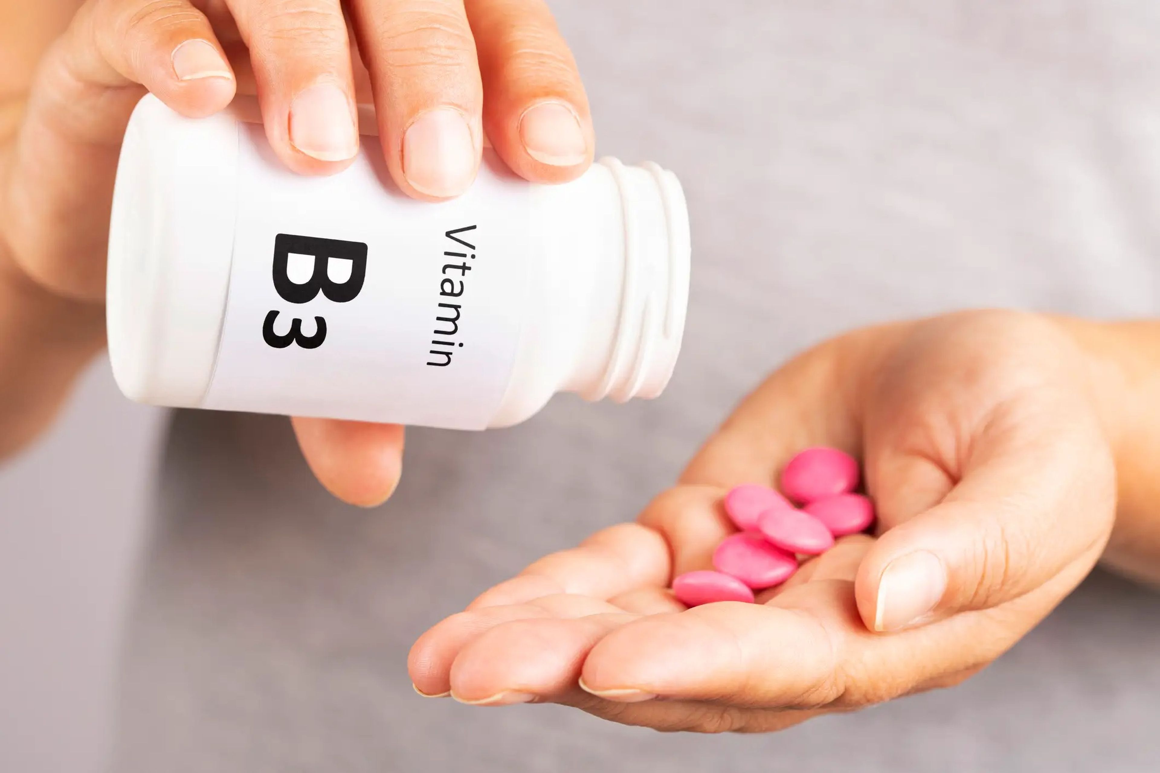 A close-up of a person's hands, one pouring pink pills from a bottle labeled "Vitamin B3" into the other hand.
